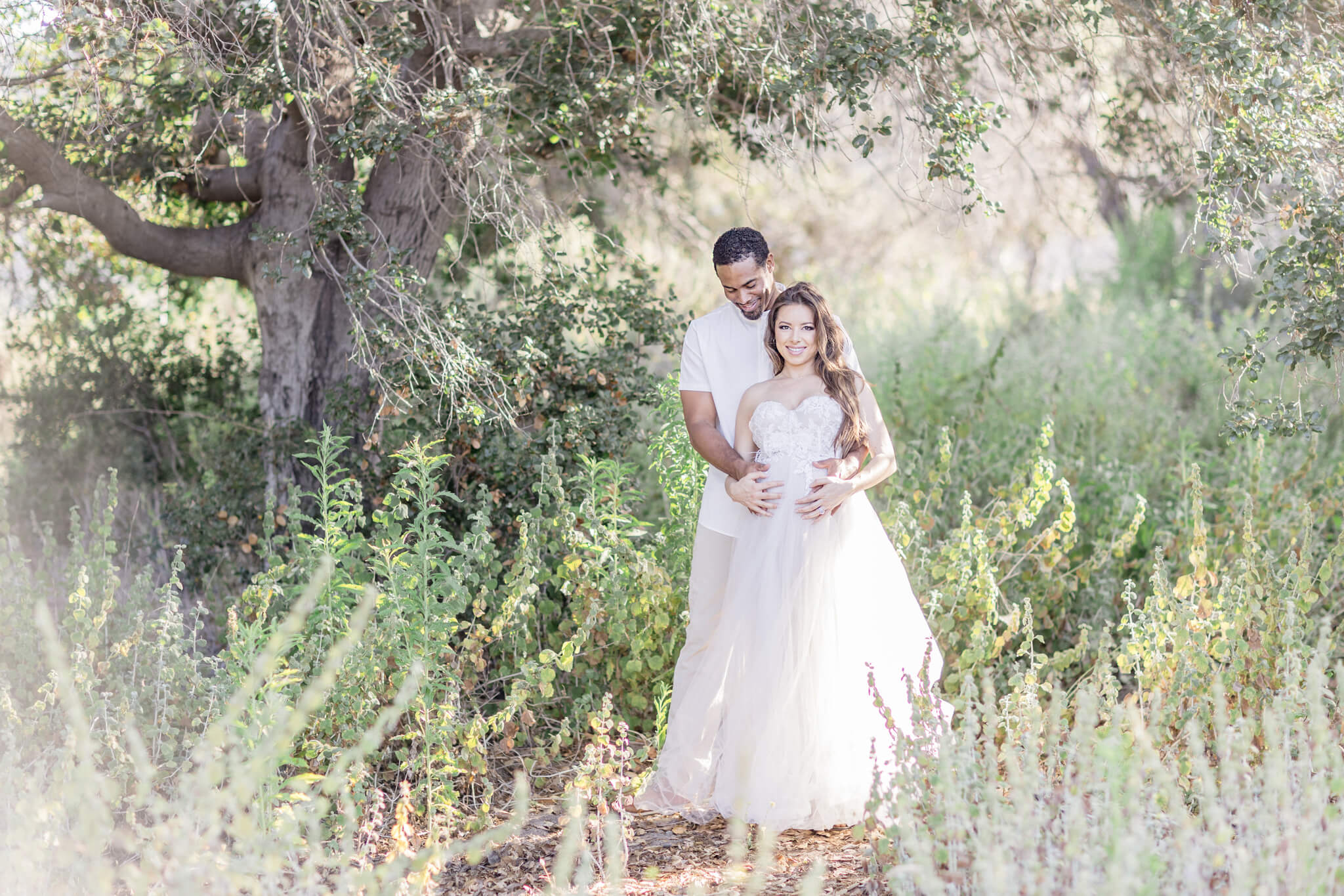 A couple stands together outdoors in a sunlit, lush green setting. The woman wears a flowing white dress, and the man stands closely behind her, holding her gently as they both smile toward the camera.
