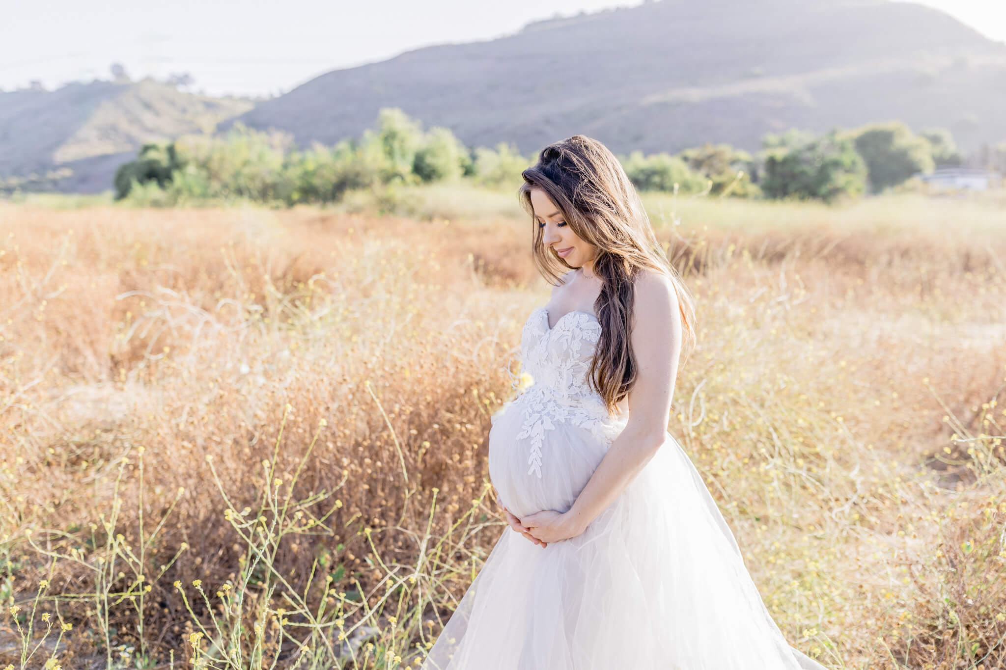 A pregnant woman in a white dress stands in a sunlit field, gently holding her belly and looking down, with hills and greenery in the background.