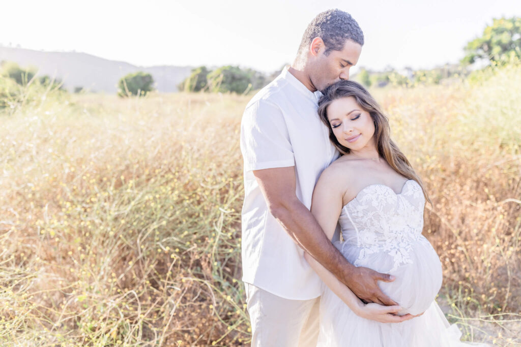 A man gently hugging a pregnant woman from behind and holding her pregnant belly in Newport Beach.