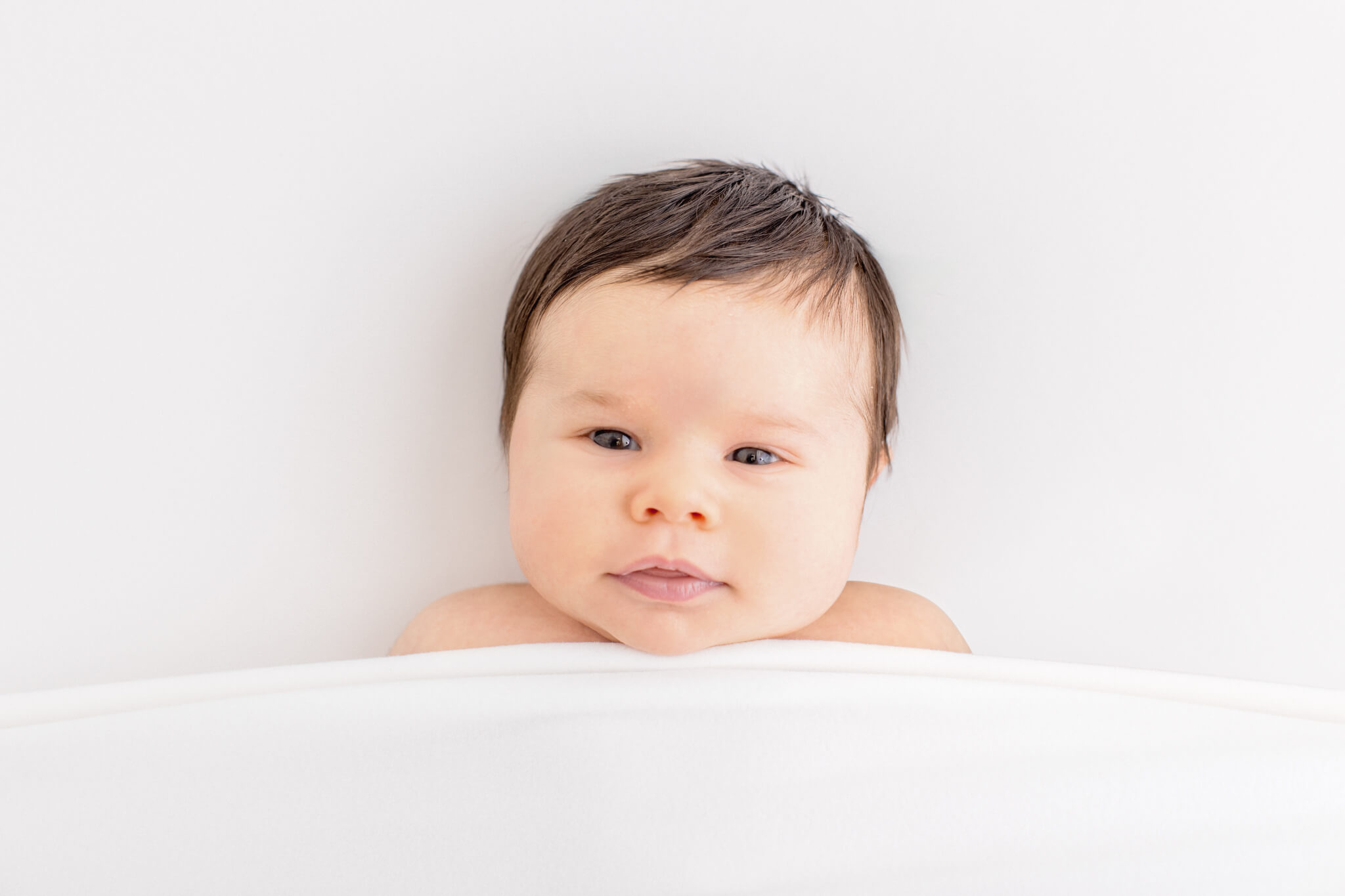 A newborn baby with black hair under a white blanket looking at the camera