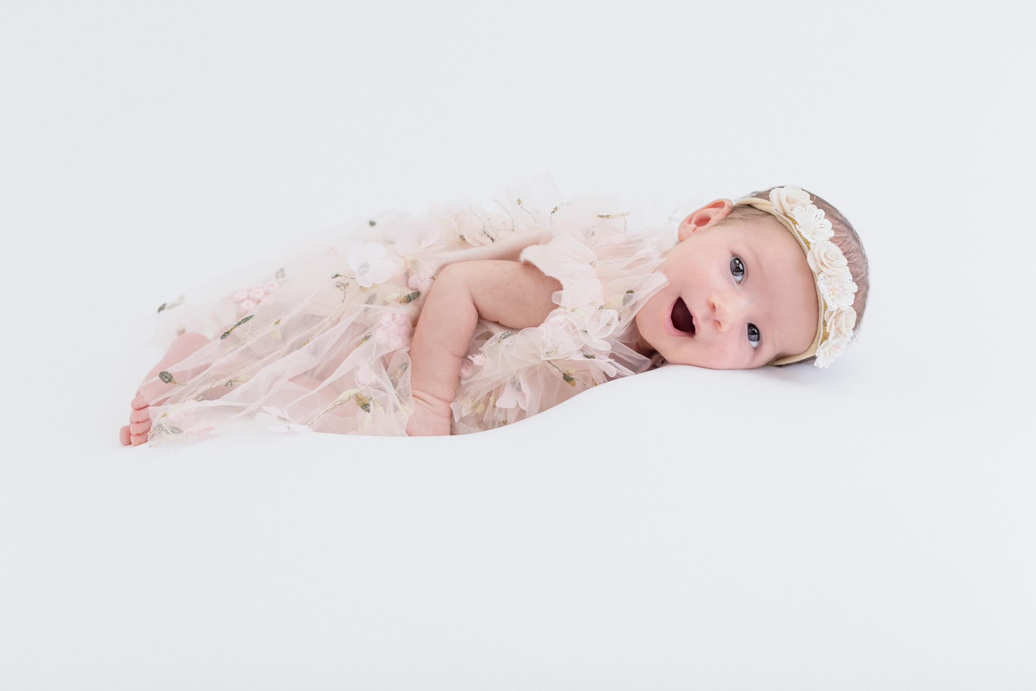 Baby in a pink floral dress laying on a white backdrop smiling at the camera