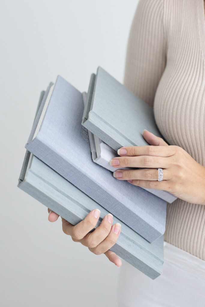 A person wearing a beige ribbed top and white skirt holds several stacked, light gray linen-covered books. The person has neatly manicured nails and wears a diamond ring on one finger.