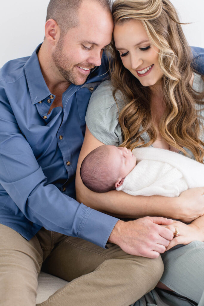 A smiling couple sits closely together, lovingly cradling a swaddled newborn baby in their arms. The mother has long wavy hair, and the father wears a blue shirt. They gaze fondly at their baby.