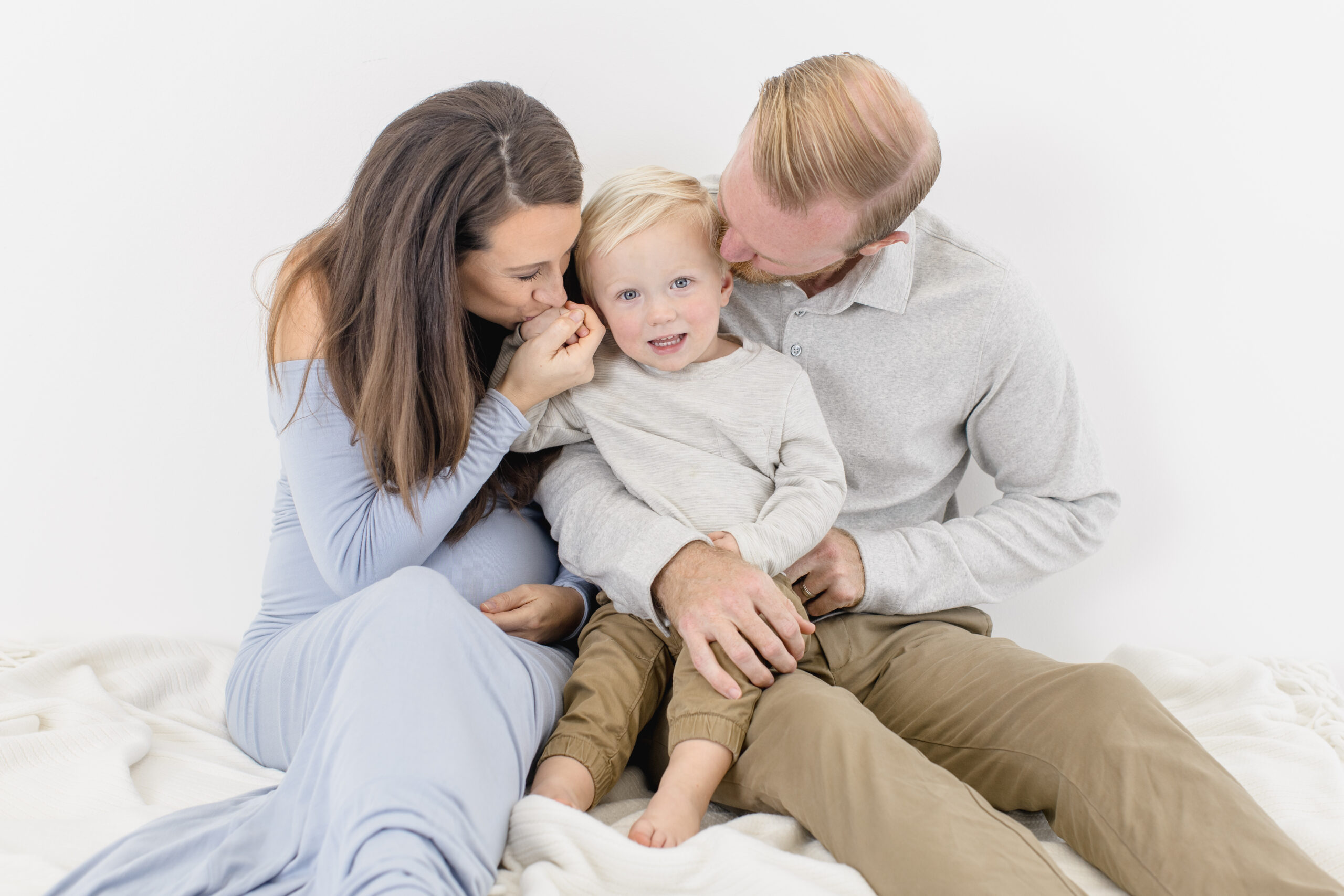 A family of three sits on a soft, white surface. The mother, in a light blue dress, and father, in a gray shirt, lovingly embrace and kiss their smiling young child seated between them.