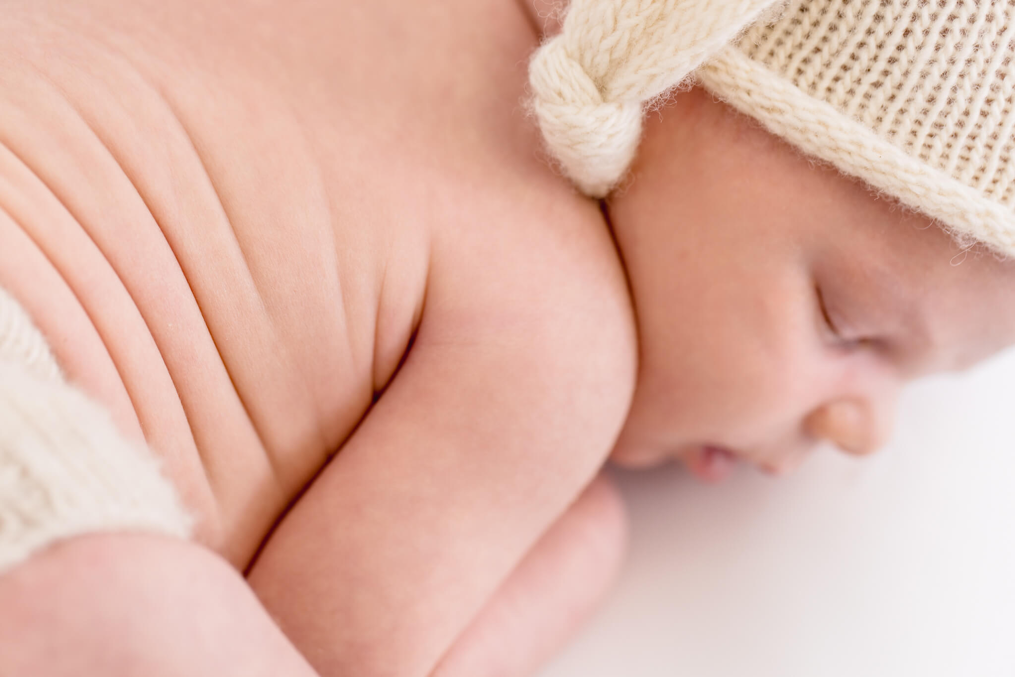 Close-up of a sleeping baby lying on its stomach. The baby is wearing a cream-colored knitted hat and matching diaper cover. Its skin appears soft with visible folds. The background is softly blurred, creating a serene and peaceful atmosphere.