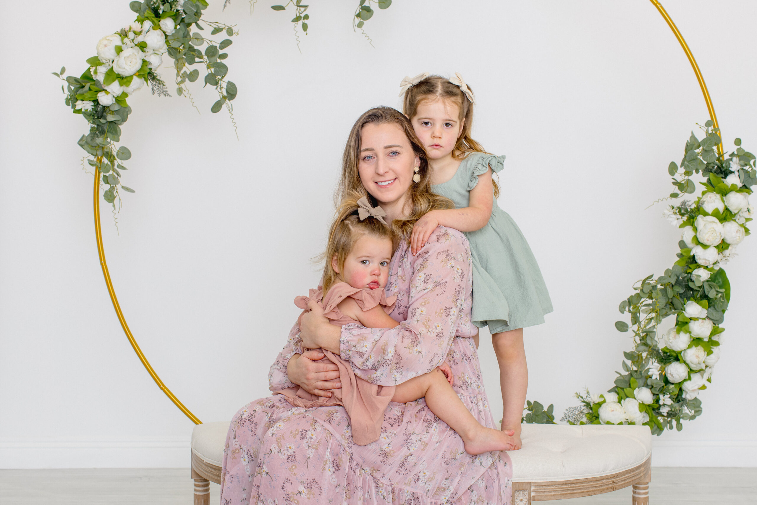 A woman in a floral dress sits on a bench, holding a young child in a pink dress, with another child in a green dress standing beside her. They are surrounded by a decorative ring adorned with green and white foliage.