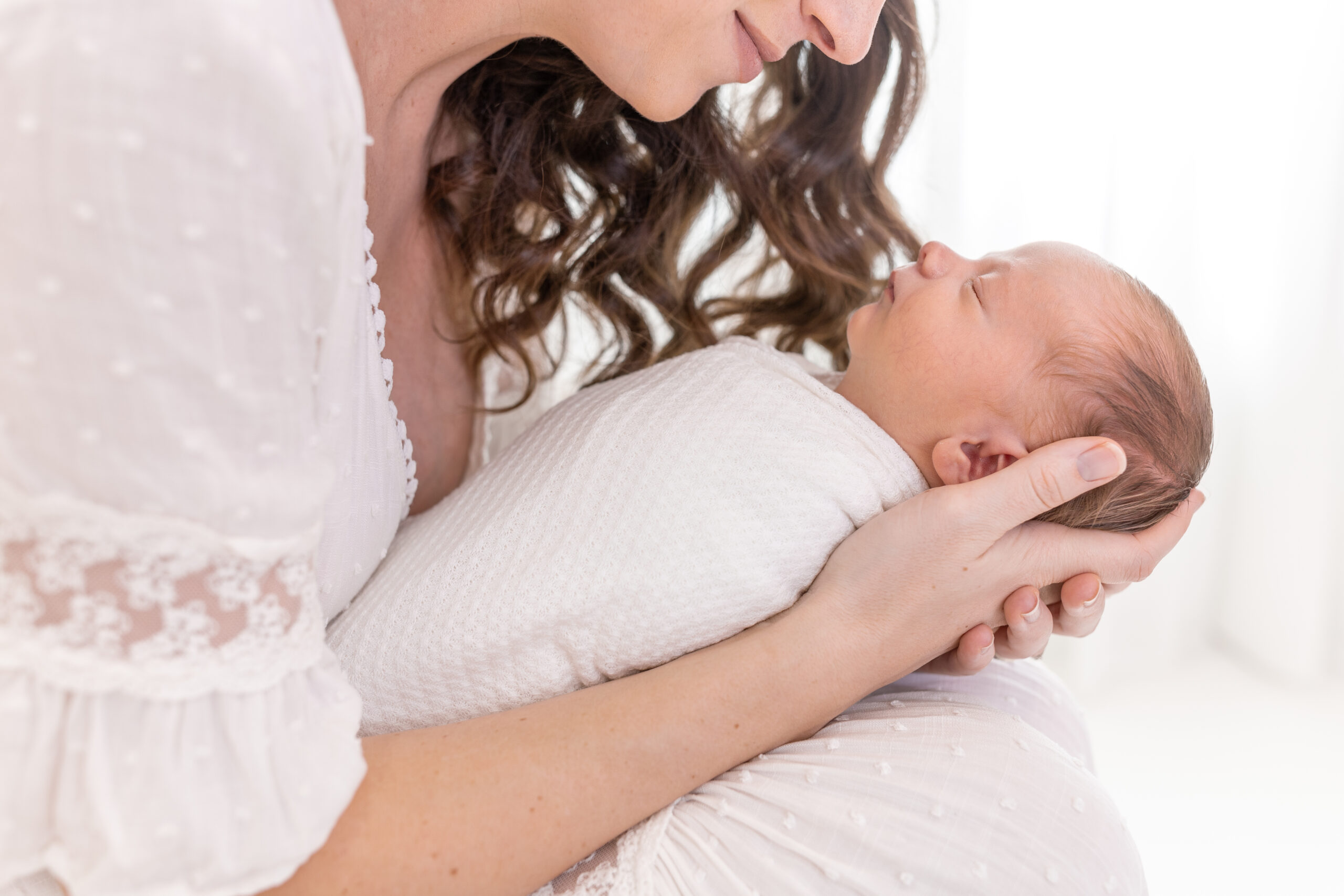 A woman gently holds a sleeping newborn wrapped in a white blanket. The scene is serene, with soft lighting highlighting their peaceful expressions. The woman has long, wavy hair and wears a textured white blouse.