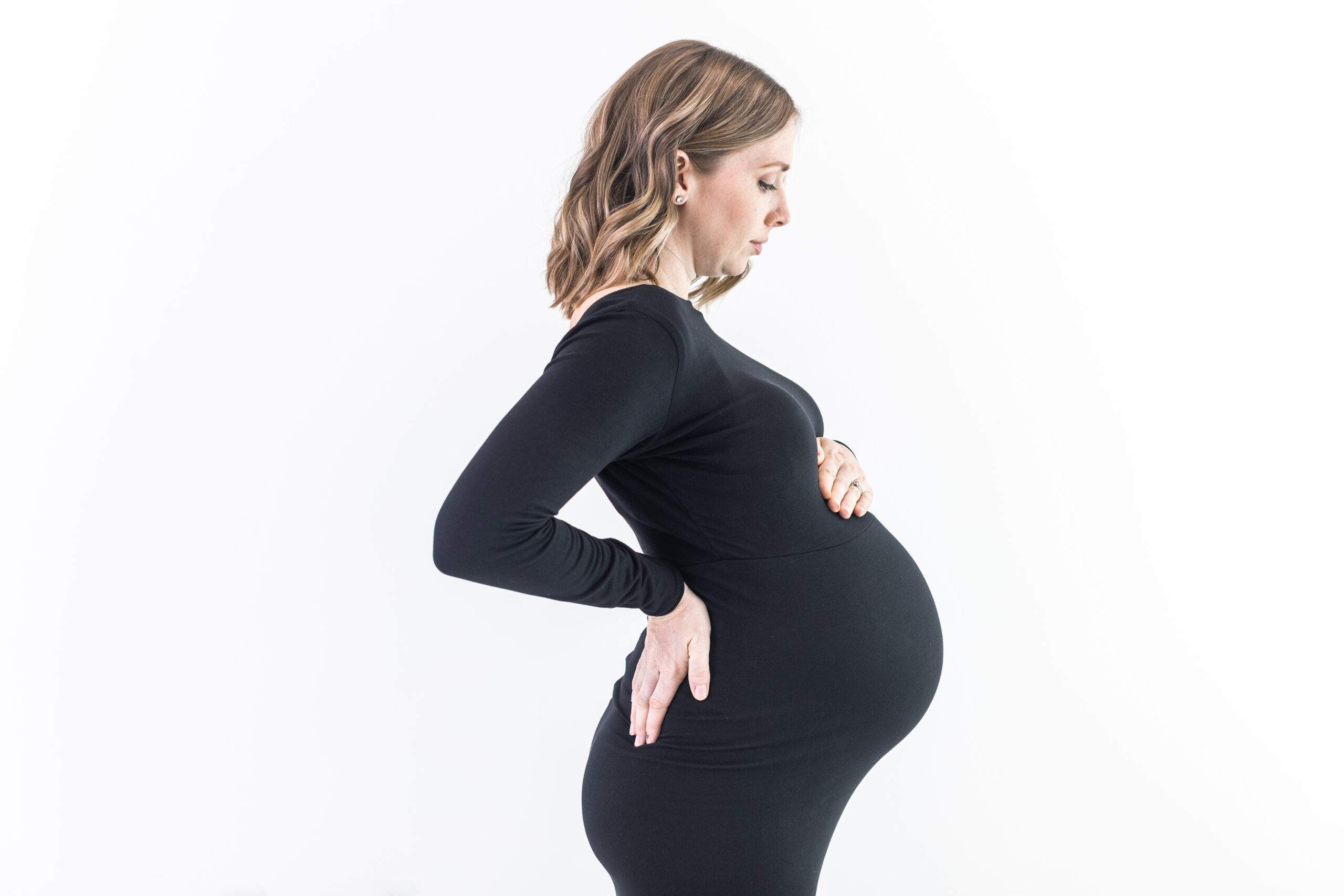 Profile of a pregnant woman standing against a white background, wearing a black long-sleeve dress. She has one hand on her lower back and the other resting below her baby bump. Her expression appears thoughtful.