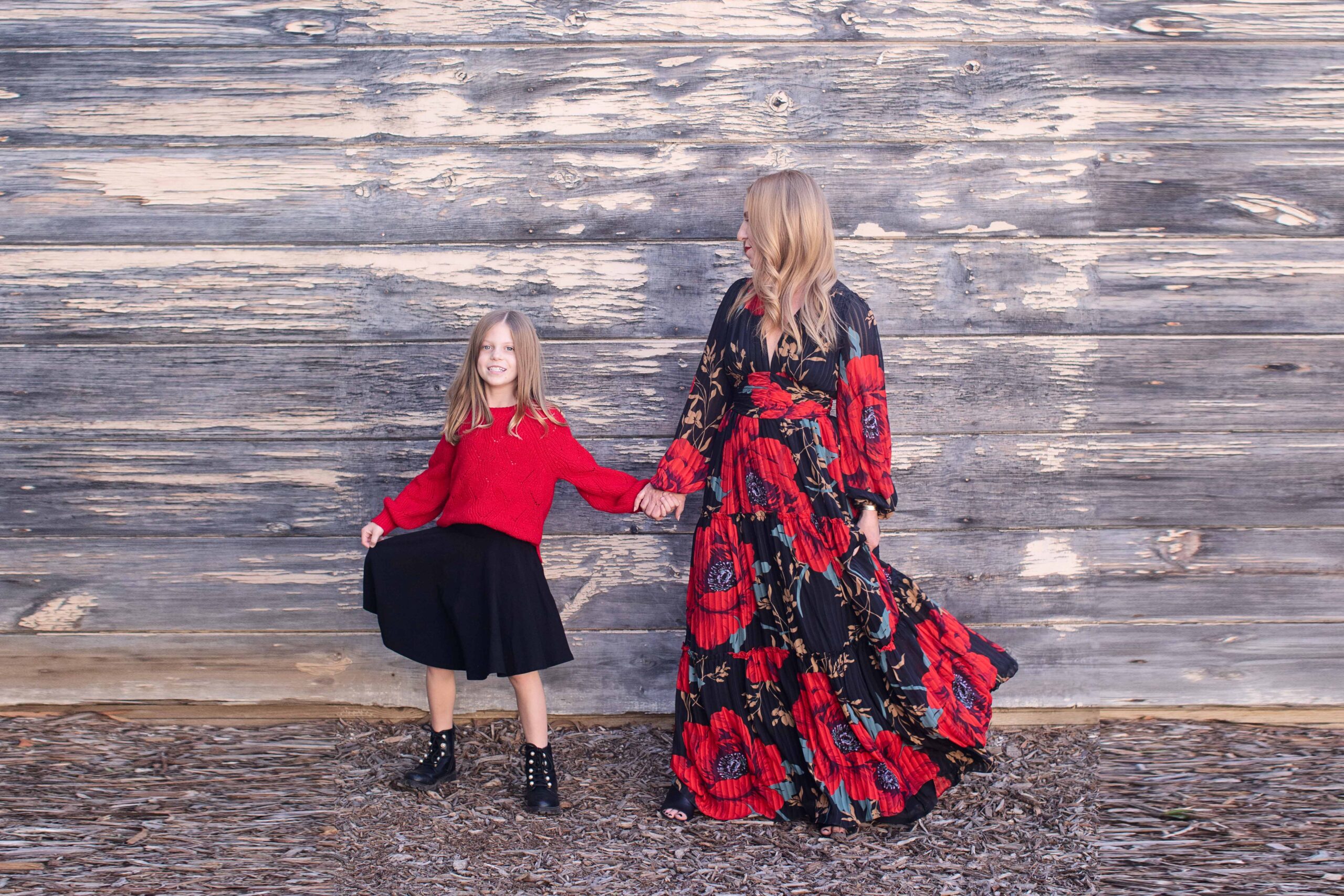 A woman in a long floral dress holds hands with a young girl wearing a red sweater and black skirt. They stand in front of a rustic wooden wall on a leaf-covered ground. The girl smiles while looking at the woman.