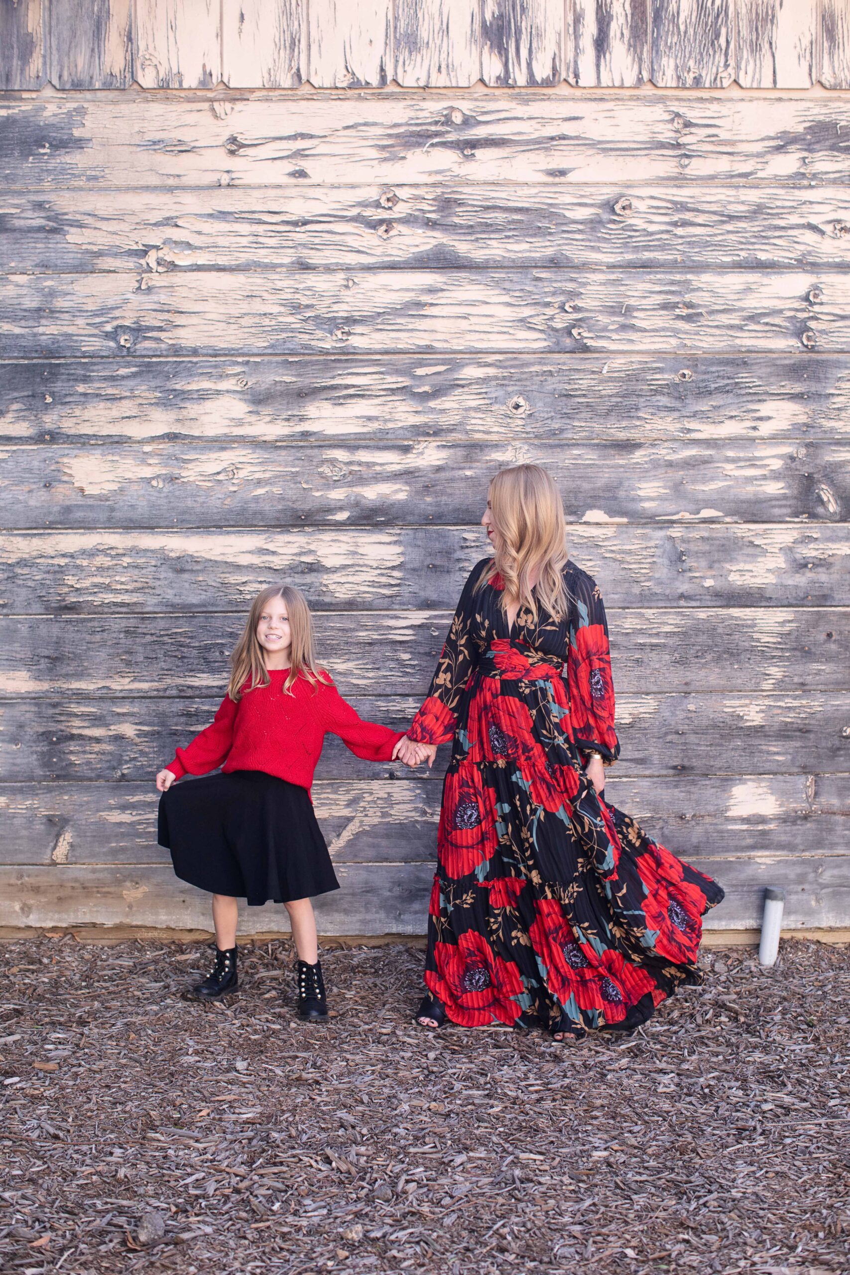 A woman and a child stand in front of a weathered wooden wall. The child is wearing a red top and black skirt, while the woman wears a flowing dress with red and black patterns. They hold hands and look slightly to the left.