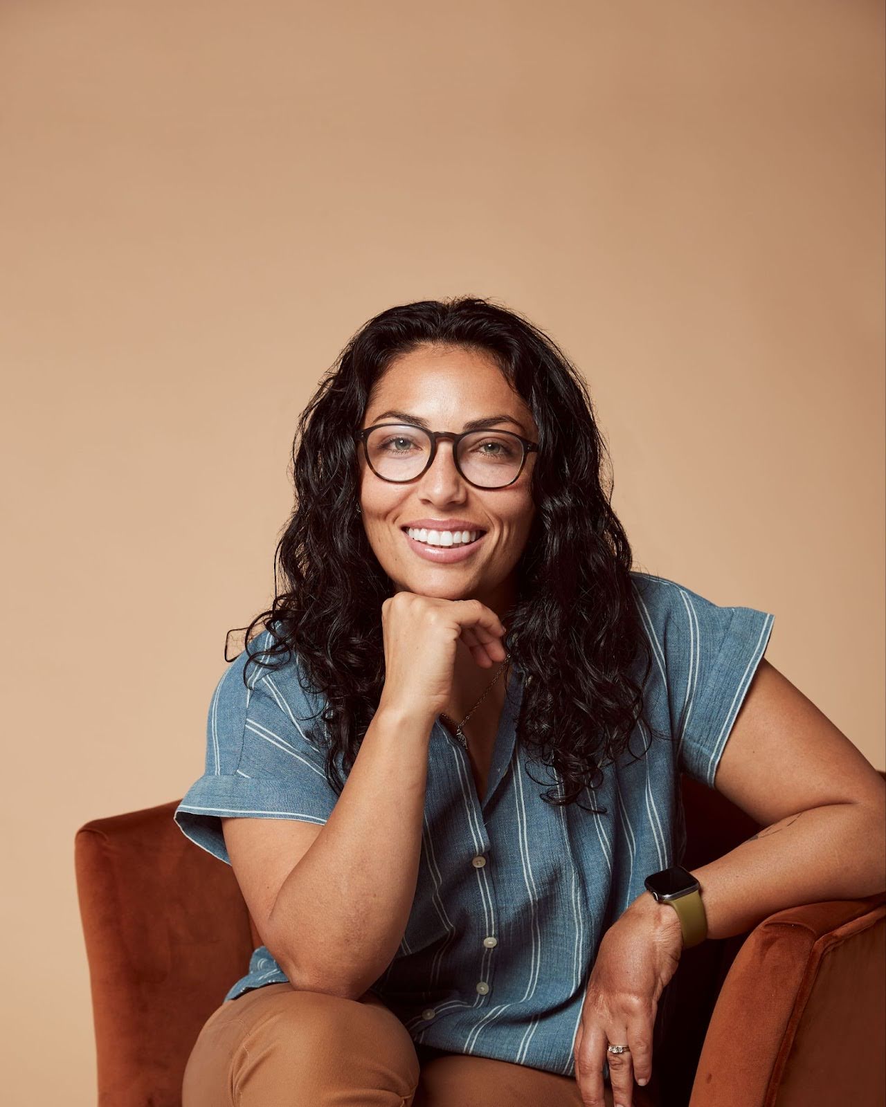 A person with long, dark curly hair and glasses is sitting on a chair against a light brown background. They are smiling, wearing a blue shirt, and resting their chin on their hand.