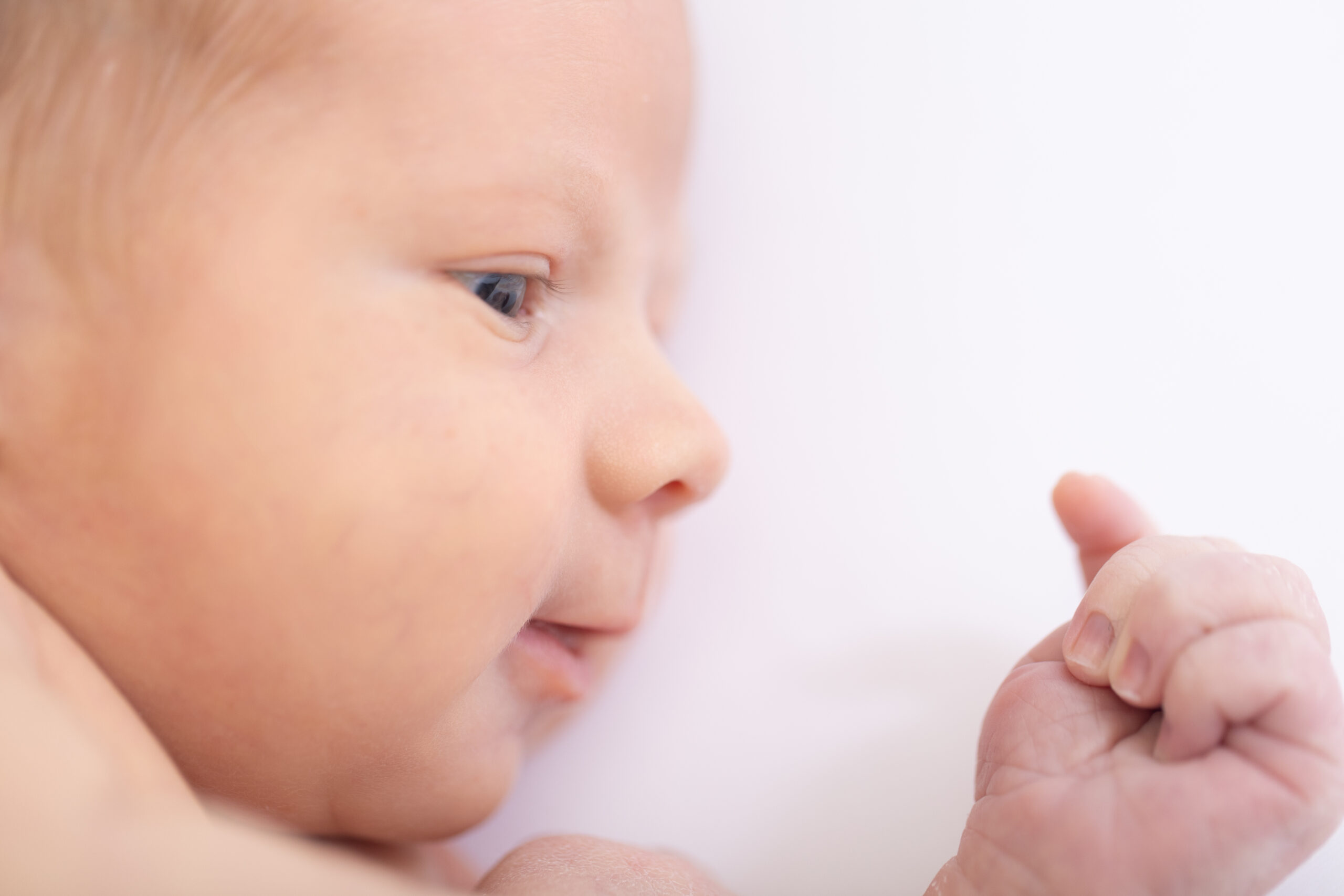 Close-up of a newborn baby lying on a white surface. The baby is looking at their hand with a curious expression. The image focuses on the babys face and hand, showcasing their delicate features and soft skin.