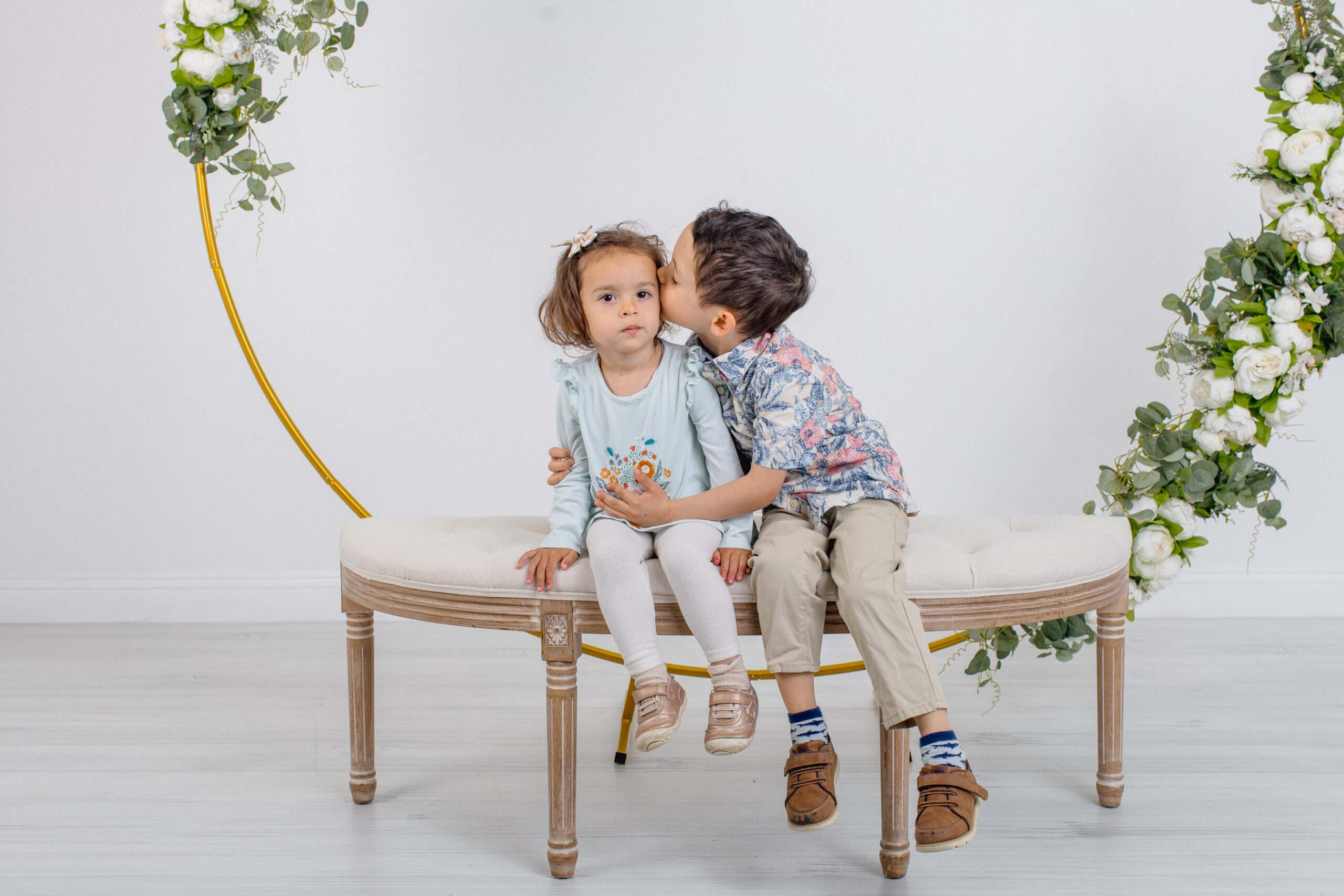 Two young children sit on a curved bench. The girl looks at the camera with a neutral expression, while the boy leans over and kisses her on the cheek. The background features a floral crescent decoration.