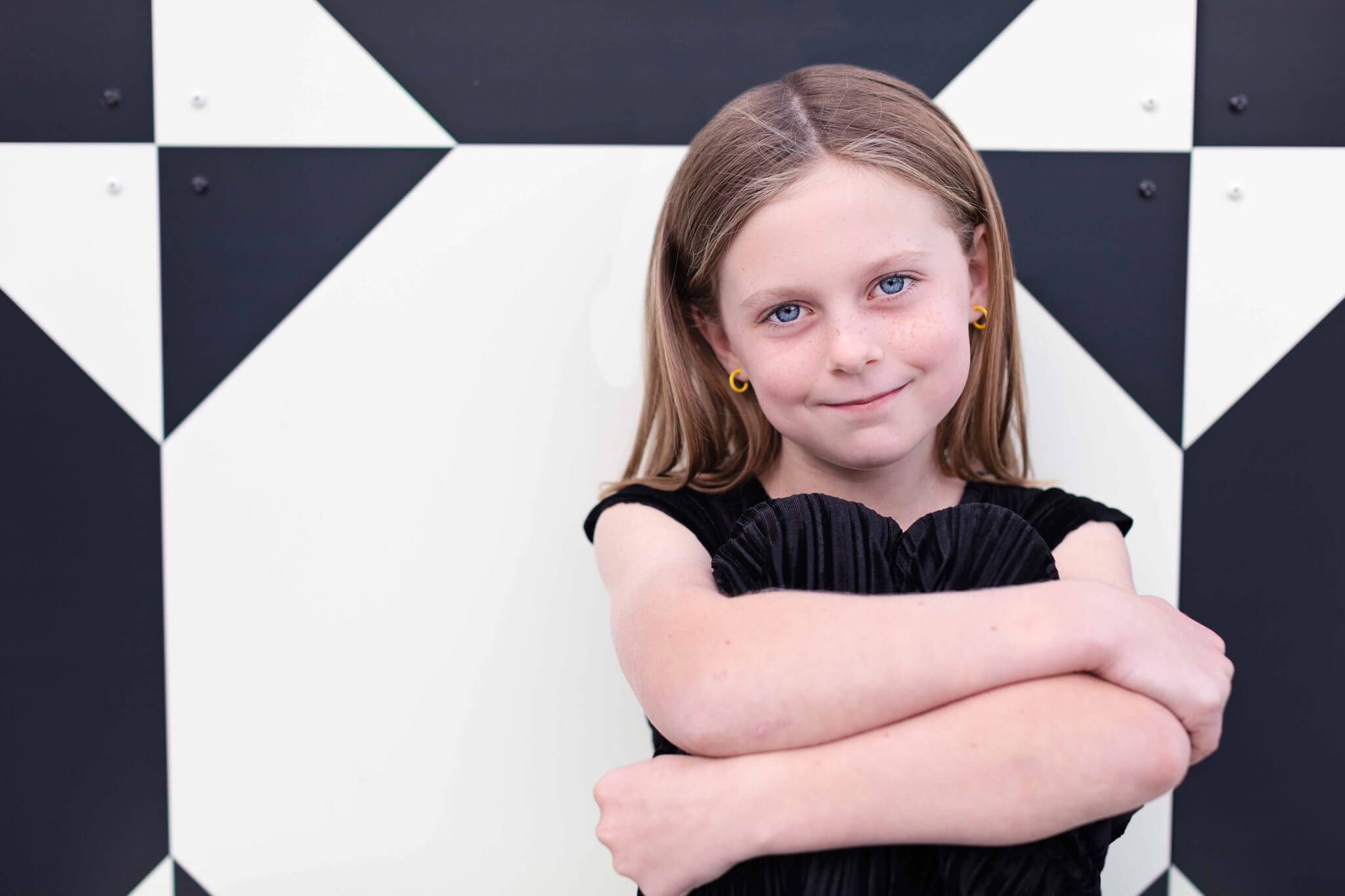 Young girl with long hair wearing a black dress, smiling and hugging her knees against a black and white geometric background.