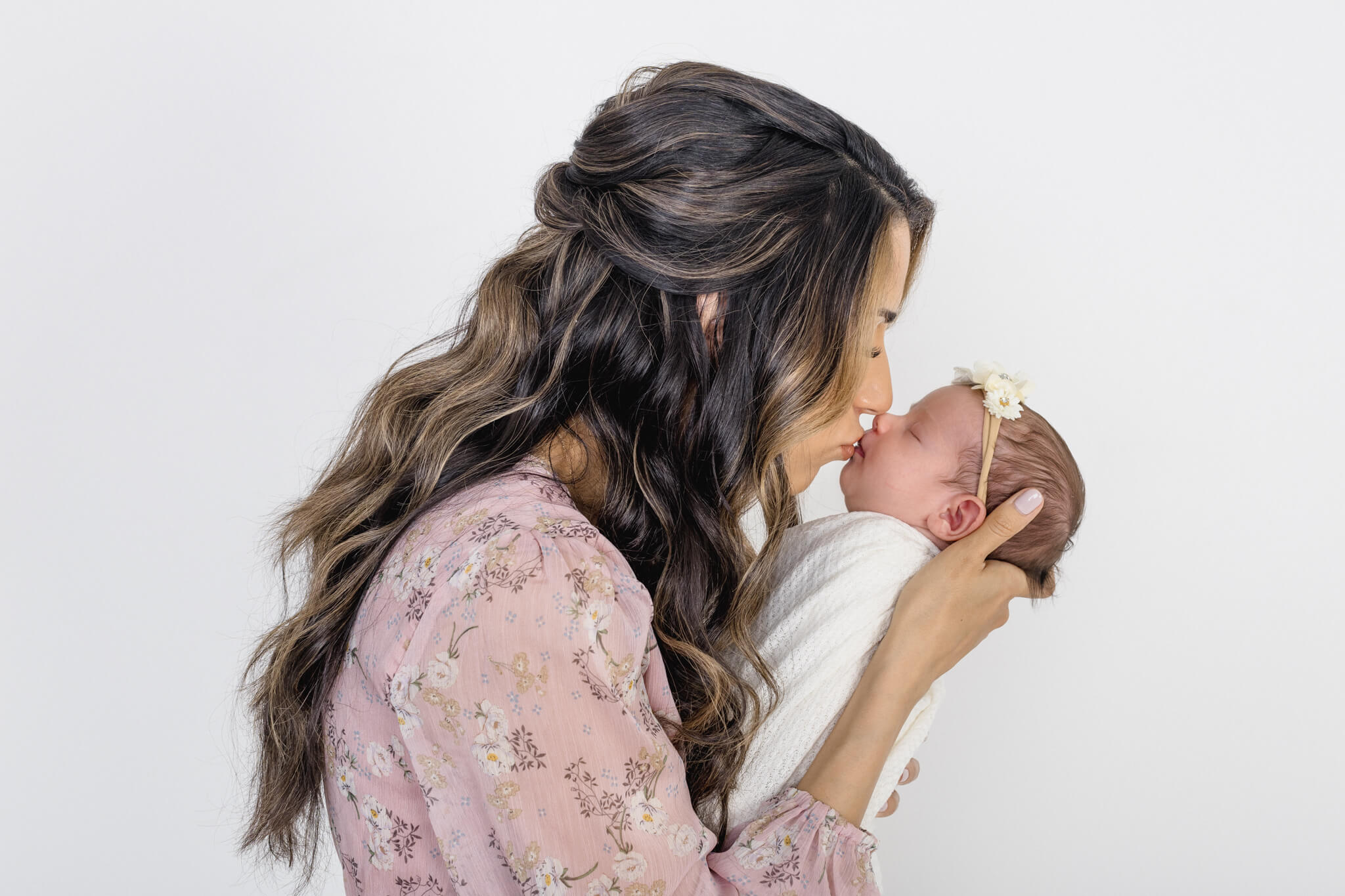 A woman with long, wavy hair kisses a newborn wrapped in a white blanket. The baby wears a delicate headband with a small flower. Both are against a plain white background, highlighting the tender moment.