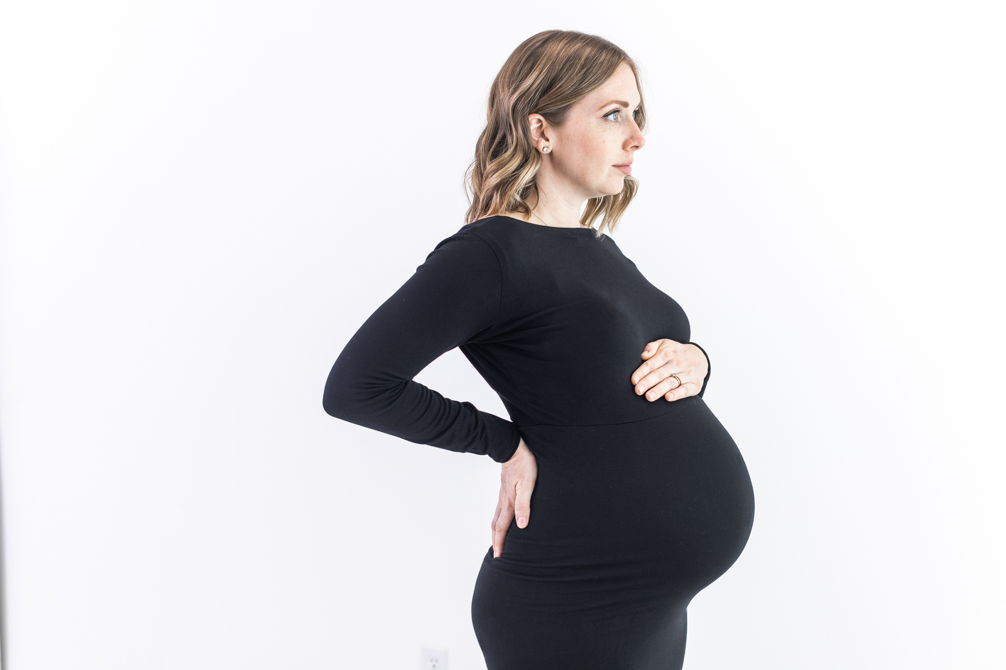 Pregnant woman in a black dress stands in profile, holding her belly with one hand and placing the other on her lower back. She is gazing to the side against a plain white background.