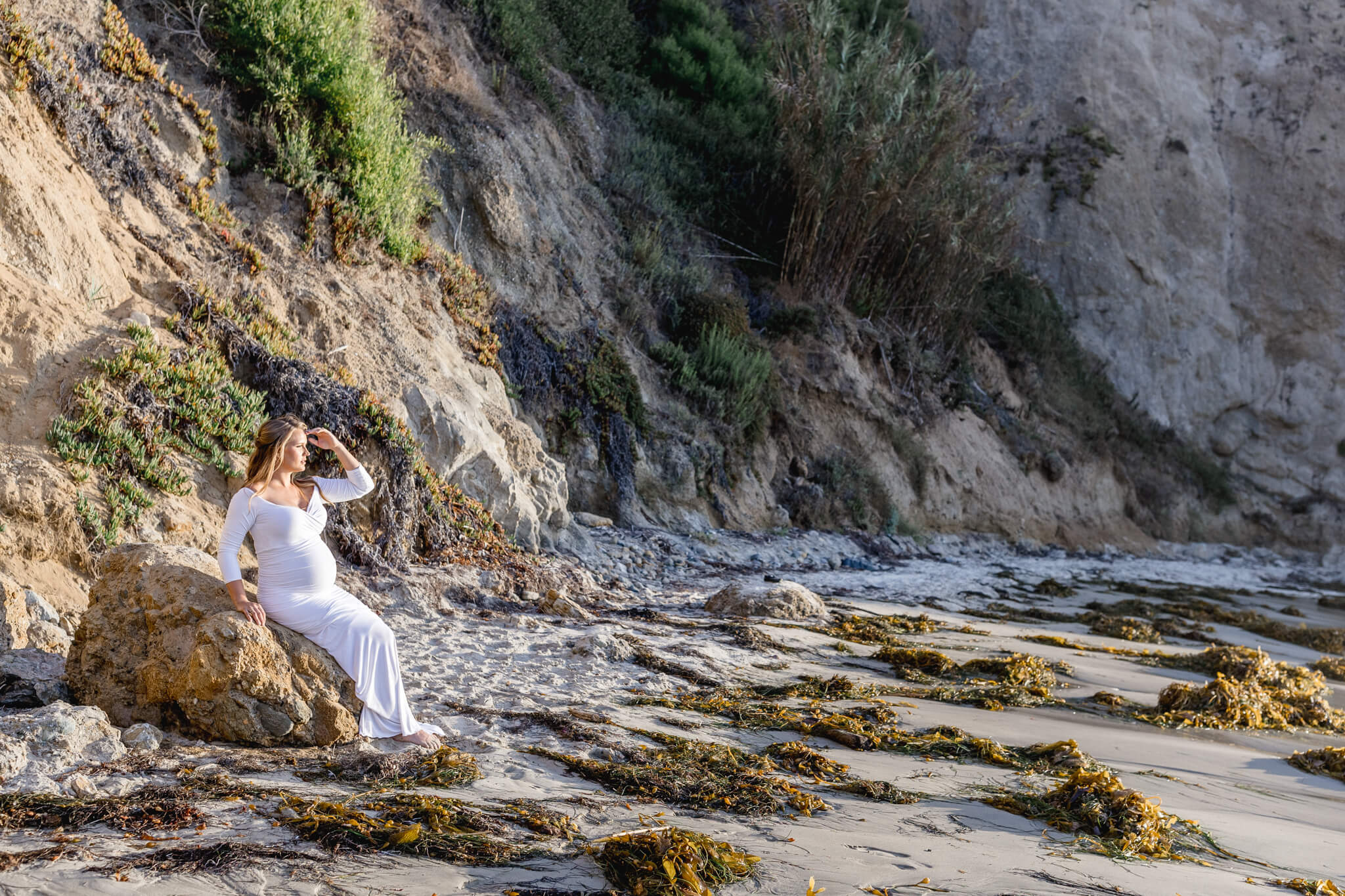A pregnant woman in a white dress sits on a large rock at the beach, surrounded by sand, seaweed, and rocky cliffs, looking into the distance with one hand shielding her eyes.