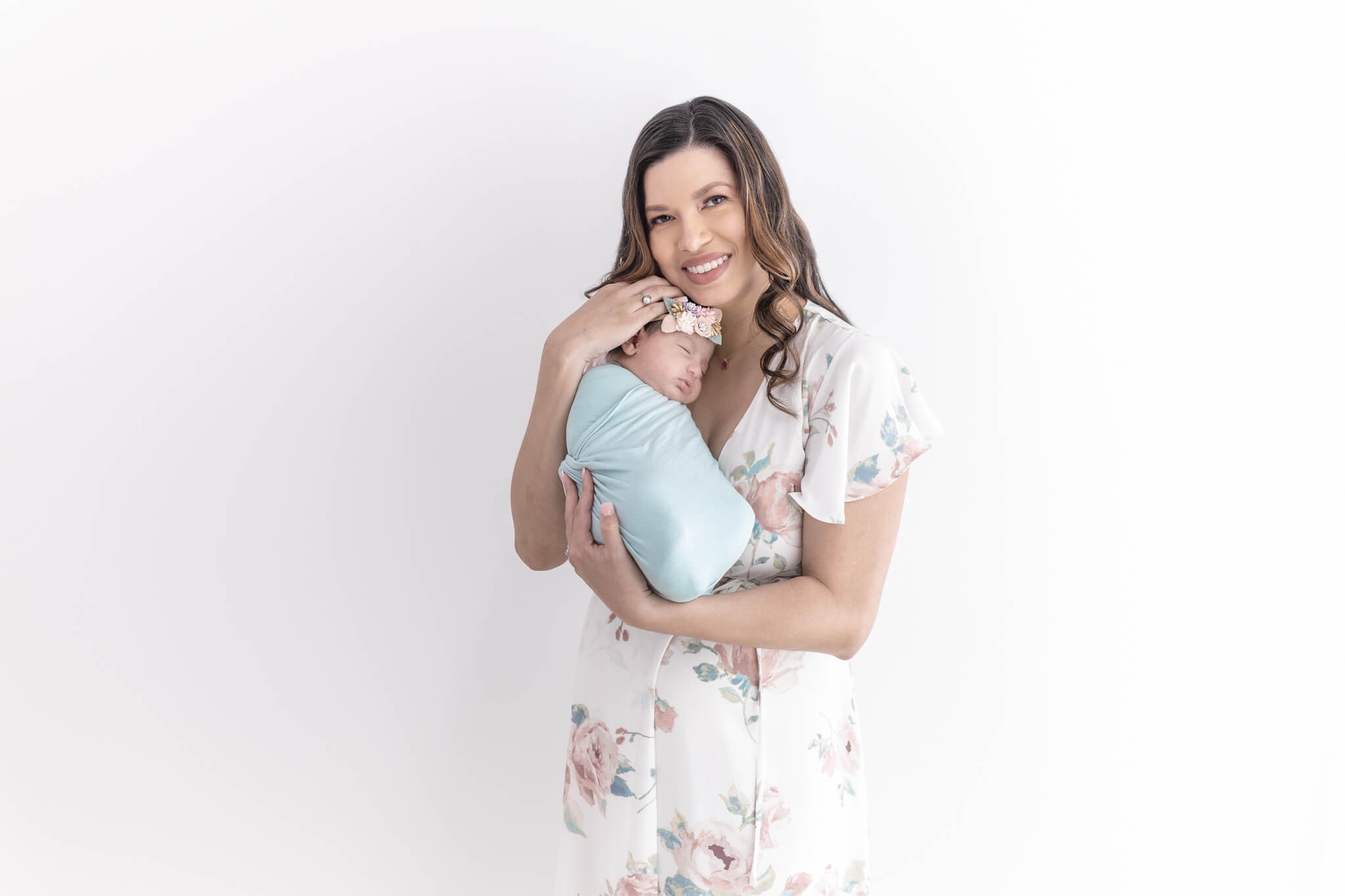 A smiling woman in a floral dress holds a swaddled newborn wrapped in a light blue blanket, gently resting her hand on the baby's head against a white background.