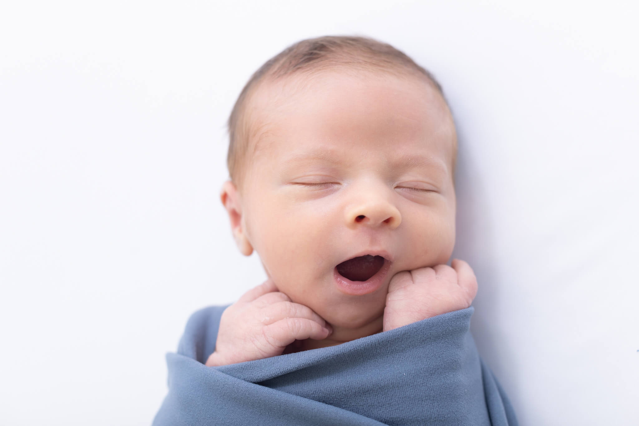 A newborn baby wrapped in a soft blue blanket lies on a white surface, eyes closed and mouth open in a yawn, with tiny hands tucked near its face.