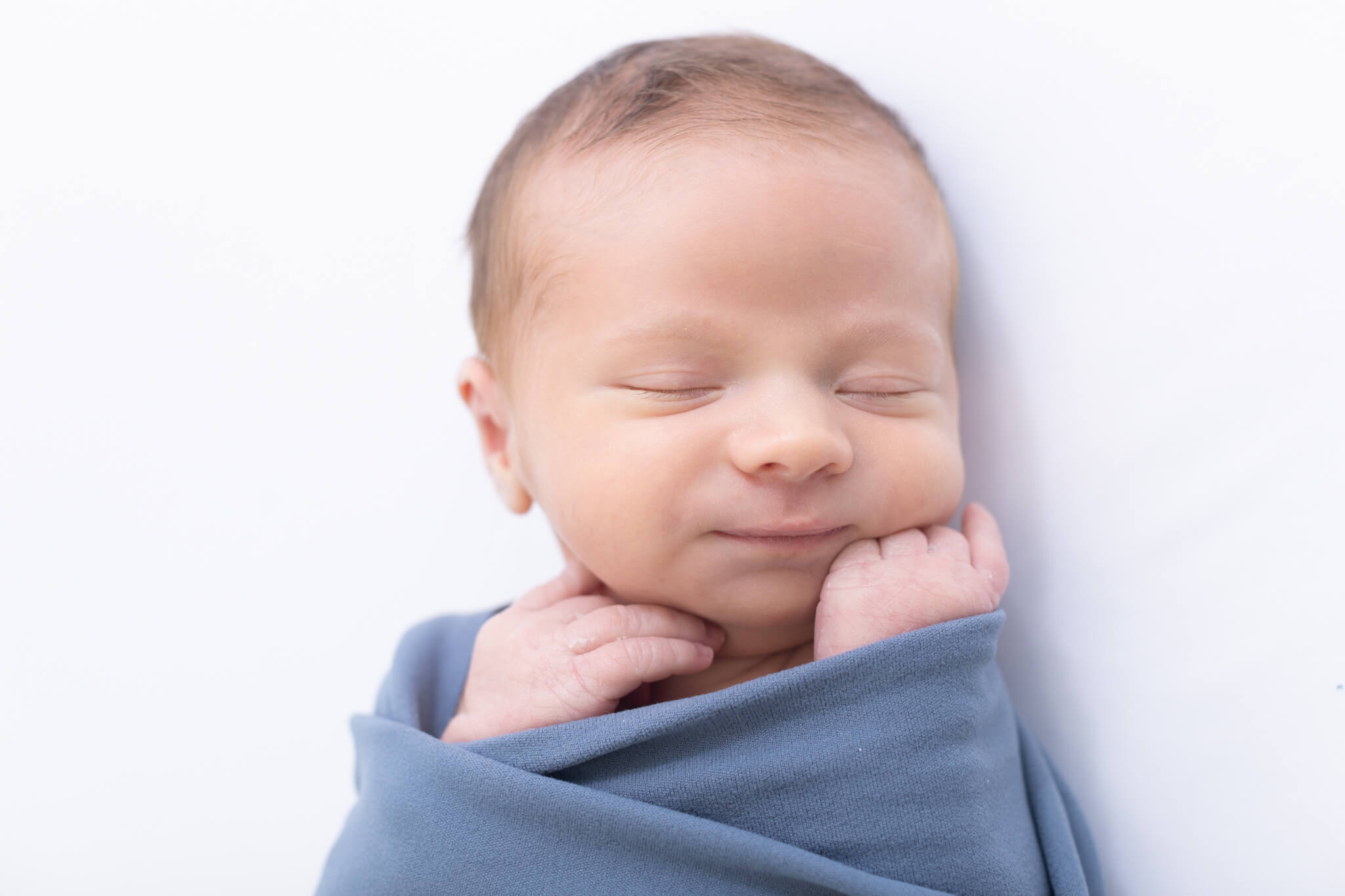 A newborn baby is wrapped in a soft blue blanket, lying on a white surface with eyes closed and a peaceful, slight smile on their face.