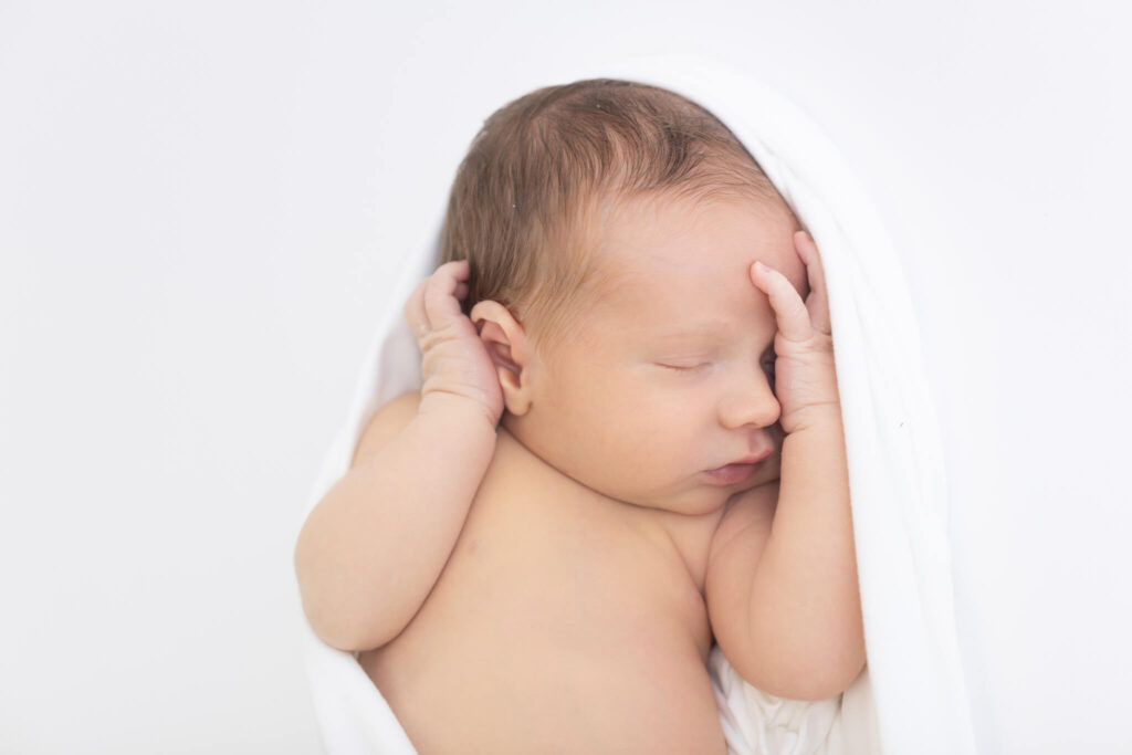 A newborn baby sleeps peacefully wrapped in a soft white blanket, with one hand resting over their face.