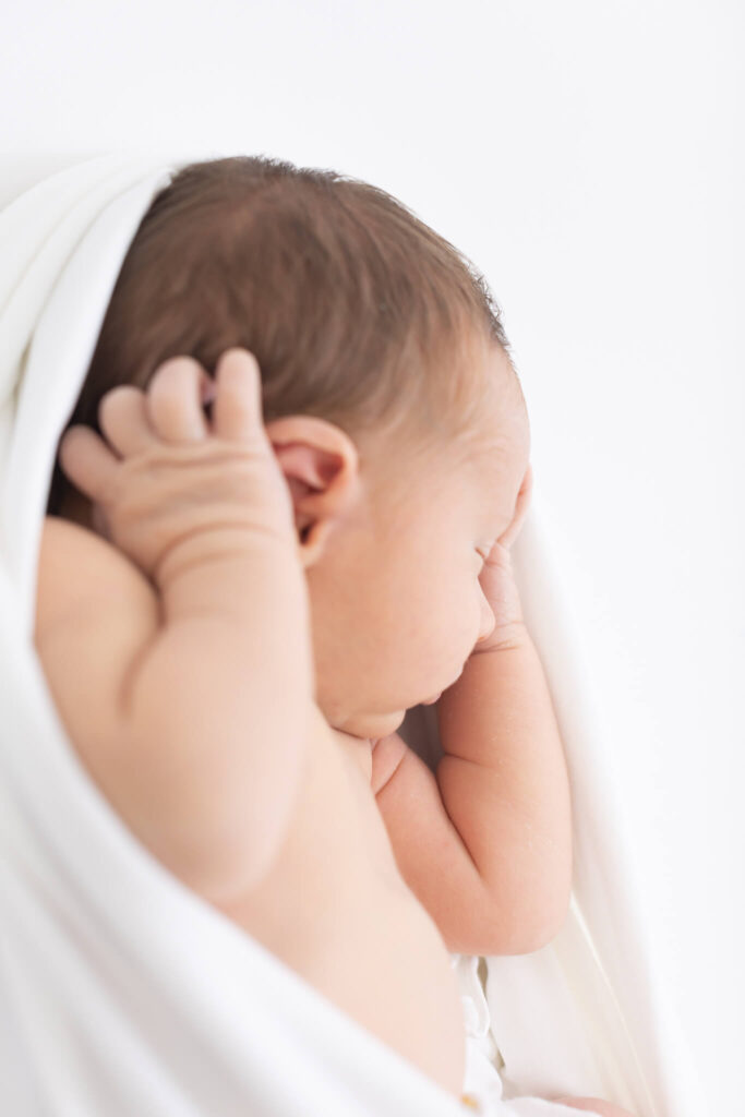 A newborn baby wrapped in a white blanket, lying on its side with hands near its face, against a soft white background.