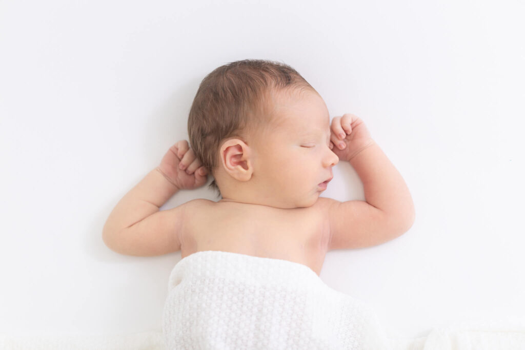 A newborn baby sleeps peacefully on a white surface, covered with a white knitted blanket, with one arm bent and the other resting above their head.