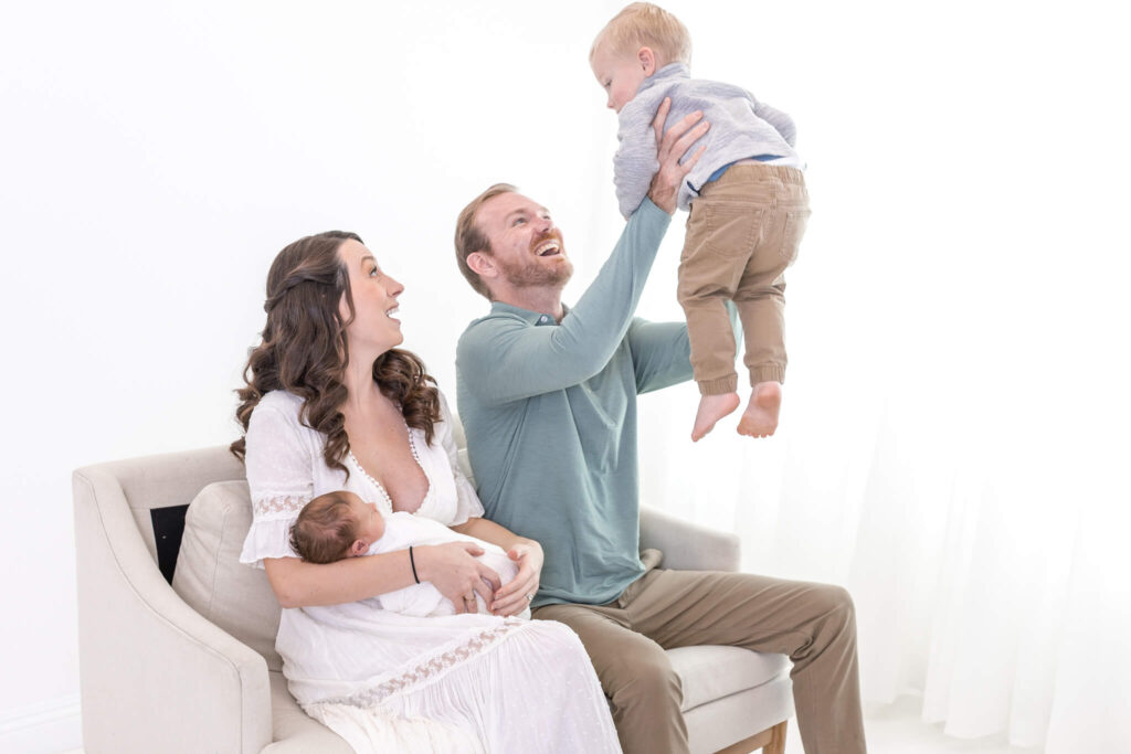 A woman in a white dress sits on a couch holding a baby, while a man beside her lifts a toddler into the air. All three adults and children are smiling and appear joyful in a bright, airy room.
