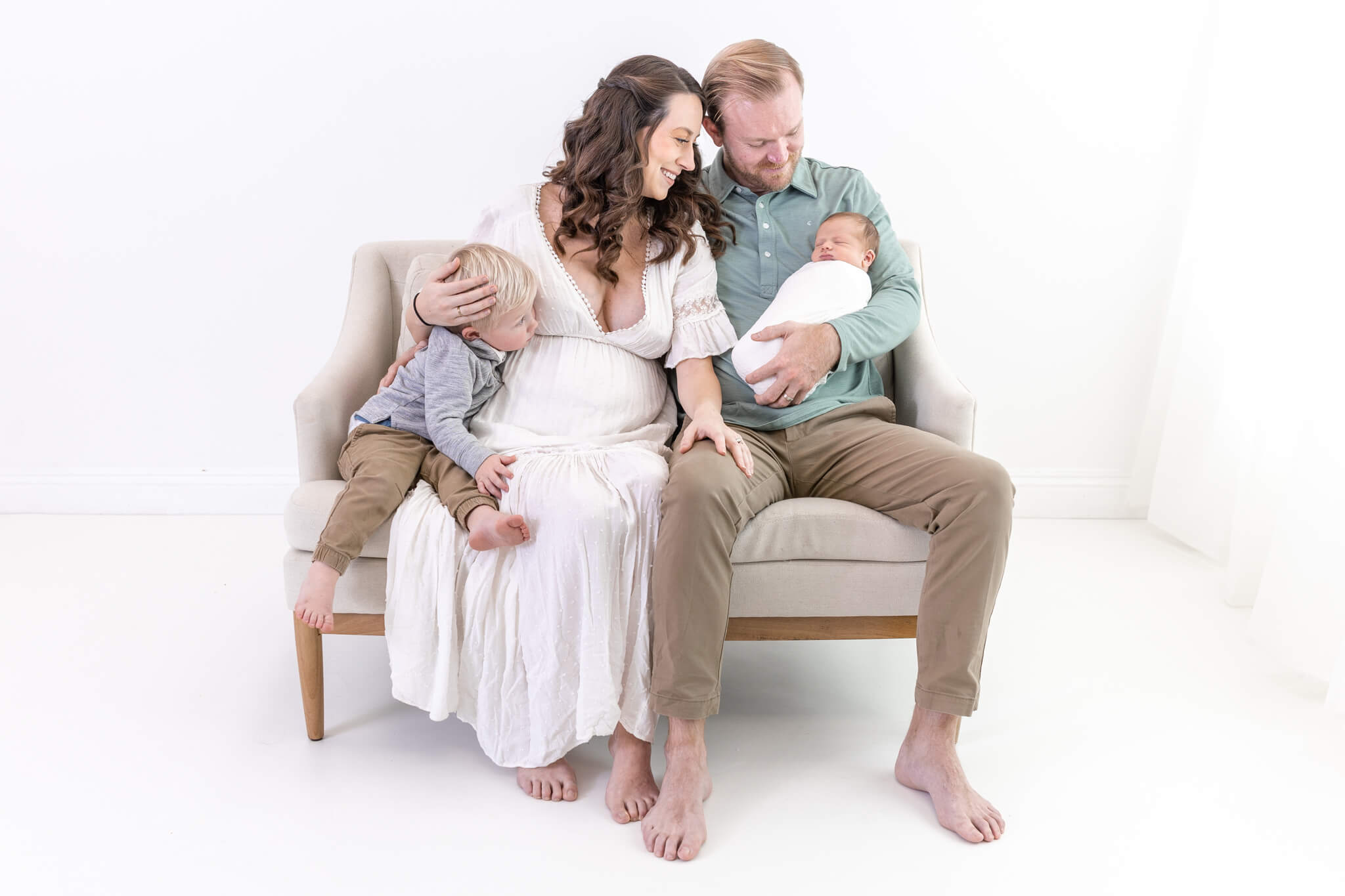 A pregnant woman in a white dress sits on a couch with a man holding a baby. A toddler sits next to her, leaning against her shoulder. All are barefoot and smiling in a bright, white studio setting.