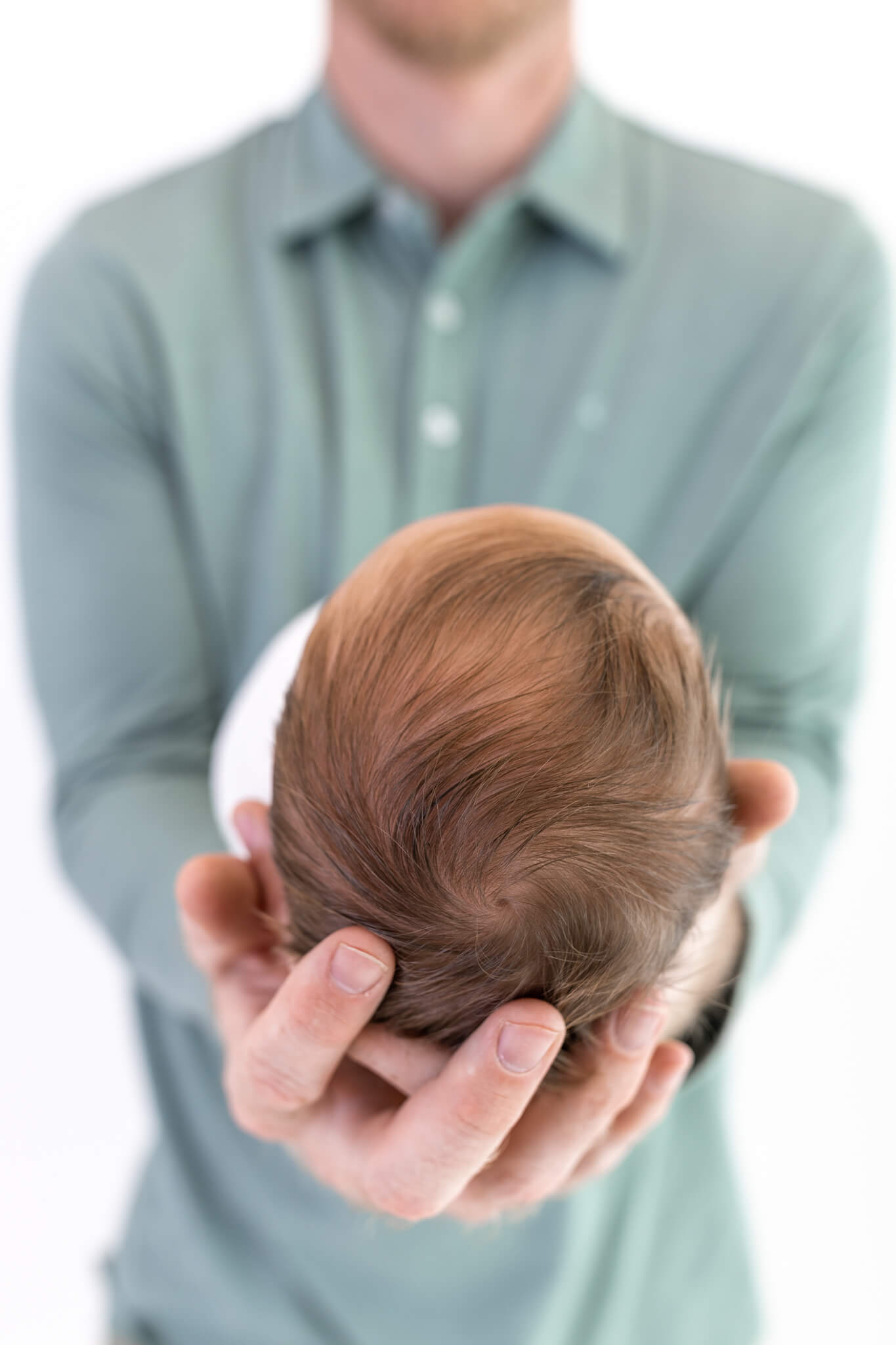 A man in a light green shirt holds a newborn baby, seen from above, cradling the babys head gently in his hands. The babys face is not visible.