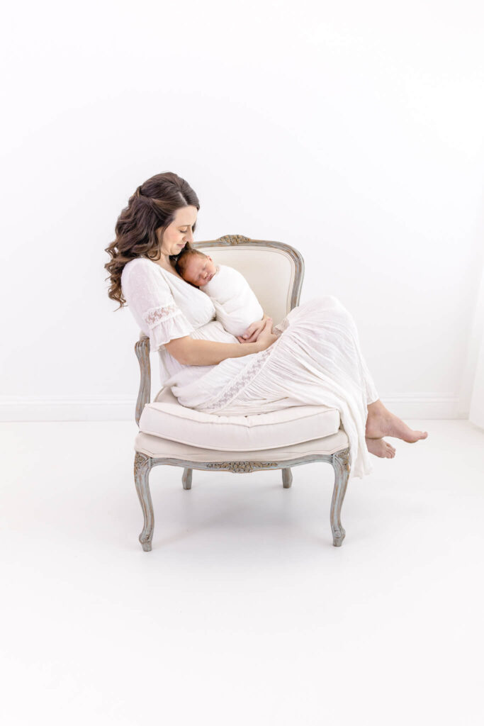 A woman in a white dress sits on an elegant chair, lovingly cradling a swaddled newborn baby in her arms against a bright, minimalistic background.