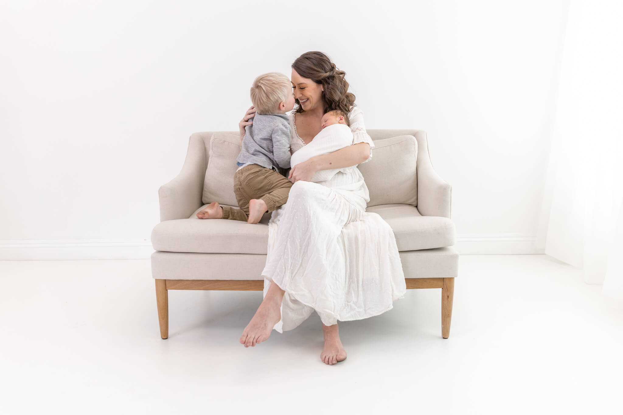 A woman in a white dress sits on a beige couch, holding a baby and smiling as a young boy in a gray shirt and khaki pants cuddles close to her in a bright, minimalistic room.