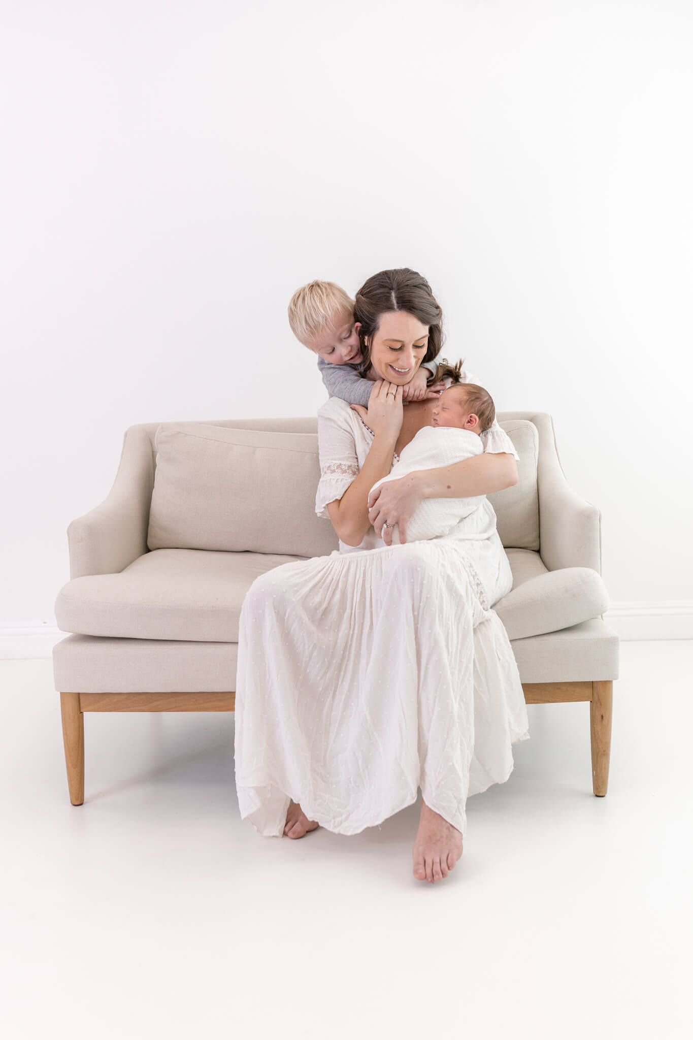 A woman in a white dress sits on a beige couch, holding a baby wrapped in white while a young child hugs her from behind. The scene is bright and softly lit, with a minimalistic white background.