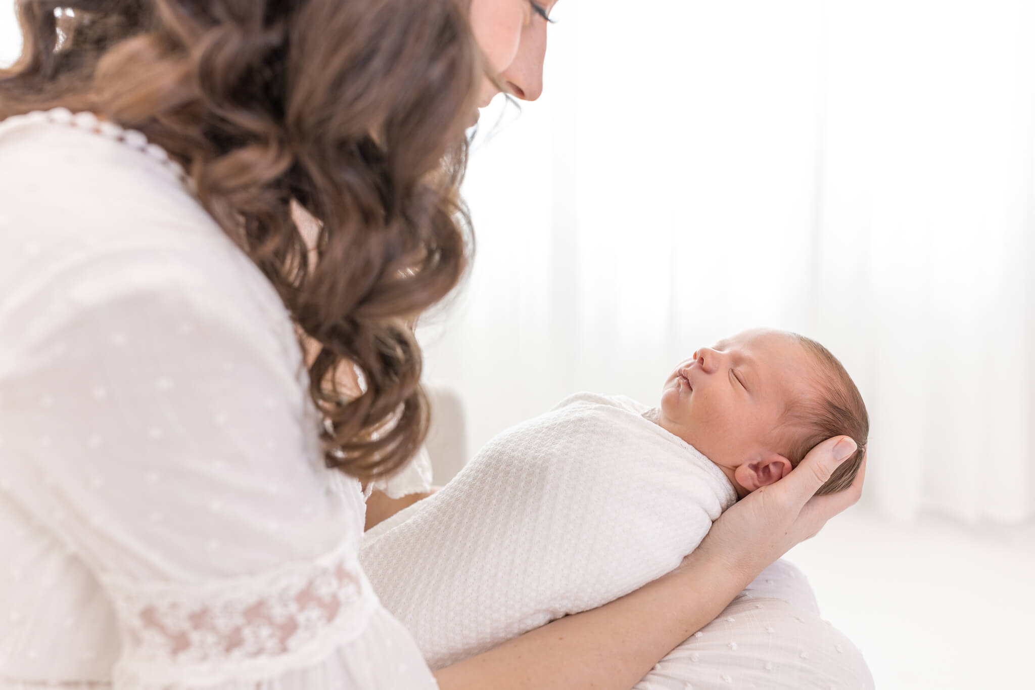  A woman with long brown hair gently cradles a swaddled newborn baby in her arms, both dressed in white, in a softly lit, bright room.