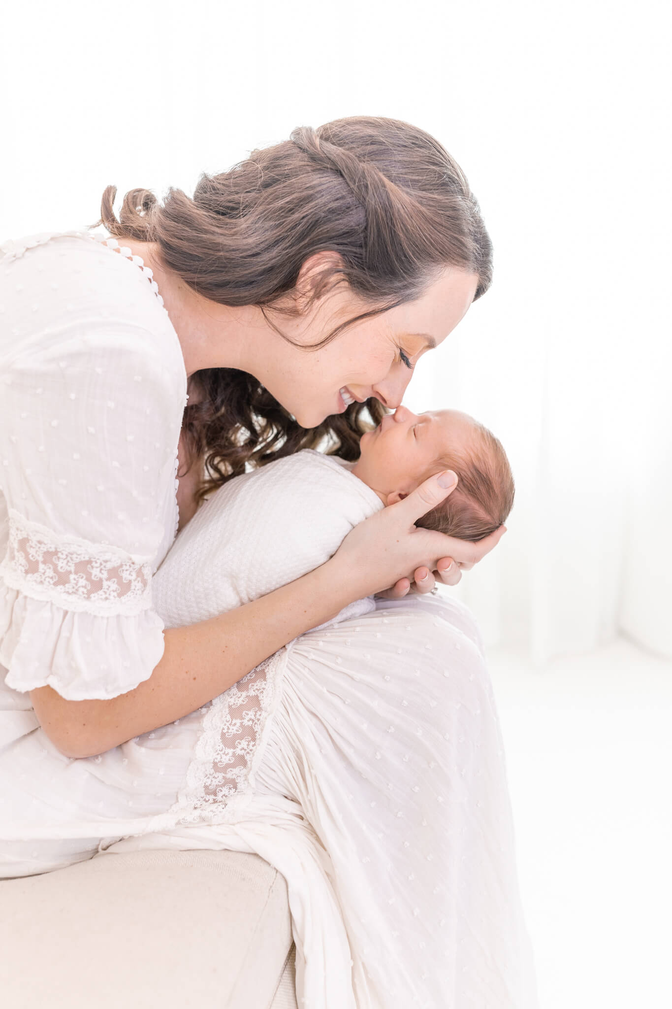 A woman in a white dress gently cradles a newborn baby, smiling as she touches her nose to the babys nose in a bright, softly lit room.