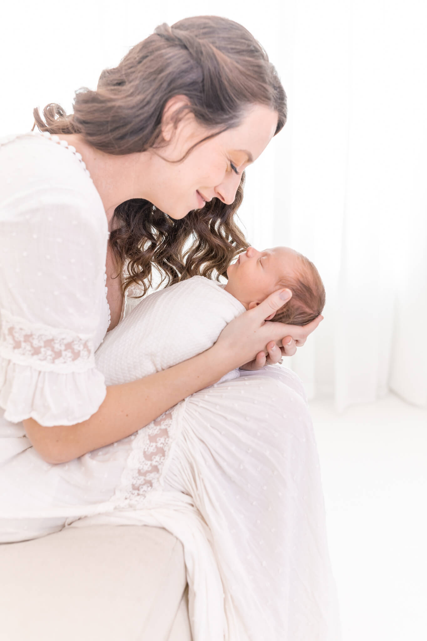a young mom in a white gauze dress holds her newborn baby boy swaddled in white in her lap and gazes down at him lovingly.