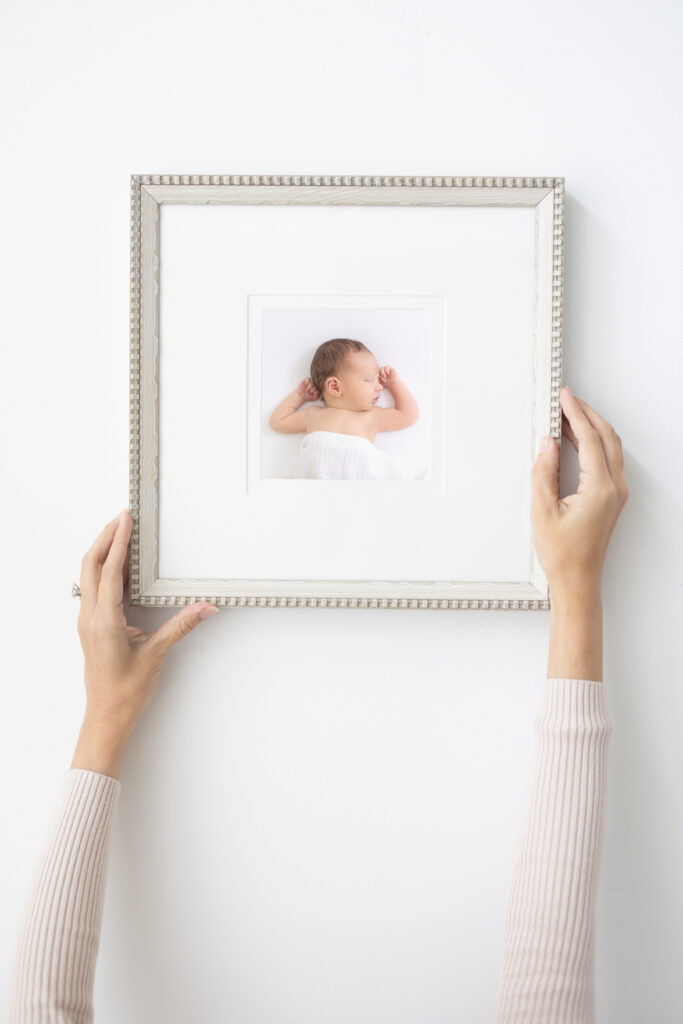 A person with light sleeves hangs a framed photo of a sleeping baby on a white wall. The baby is lying on its back with one arm raised, and the frame is square with a decorative border.
