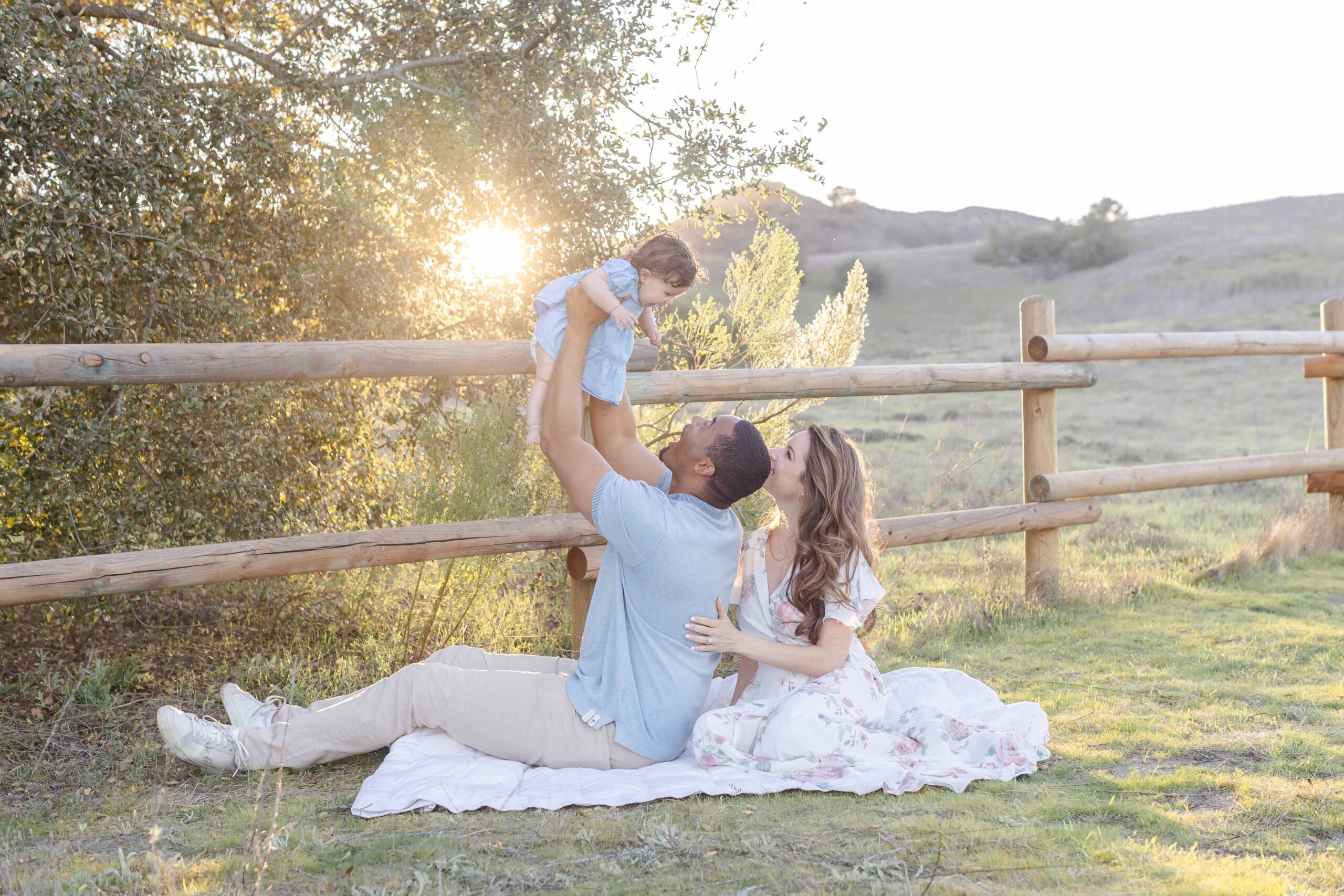 A man and woman sit on a blanket outdoors near a wooden fence. The man lifts a baby into the air as the woman looks on, smiling. Sunlight filters through the trees, creating a warm, peaceful scene.