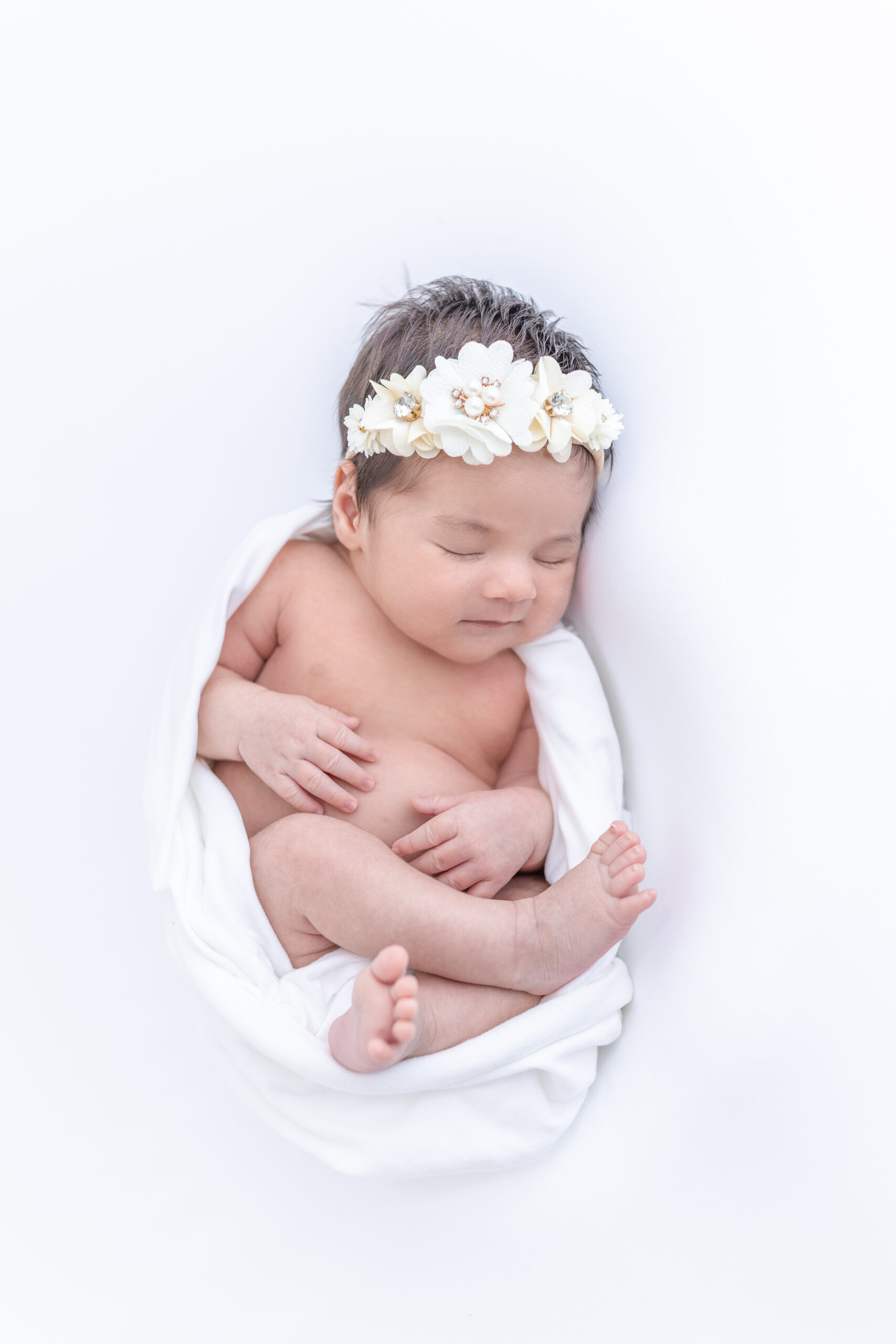 Newborn baby wrapped in a white blanket, lying on a white background, wearing a white floral headband, peacefully sleeping with hands and feet visible.