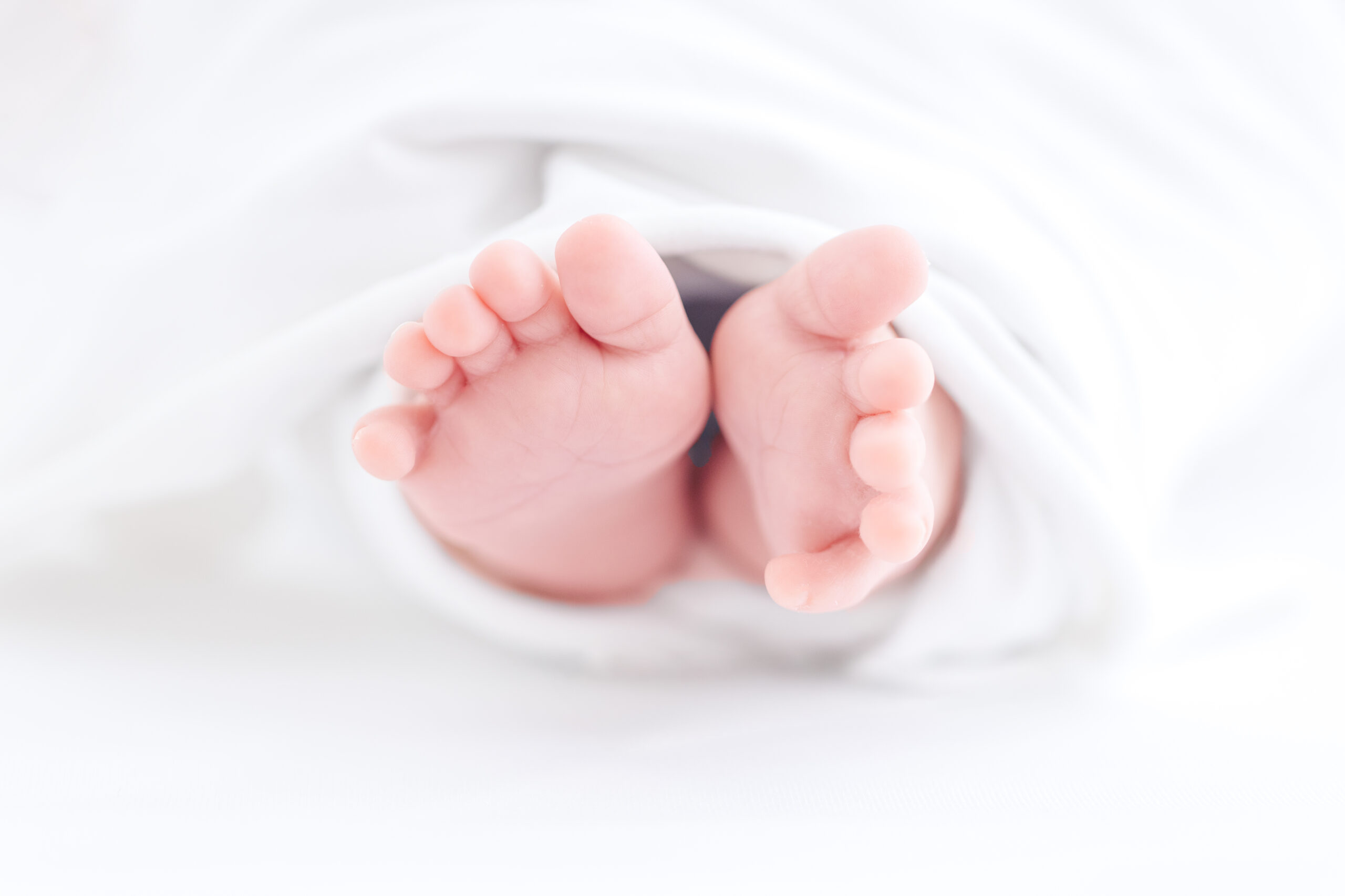 Close-up of a newborn babys feet wrapped in a soft white blanket, with toes peeking out and the background blurred in white tones.