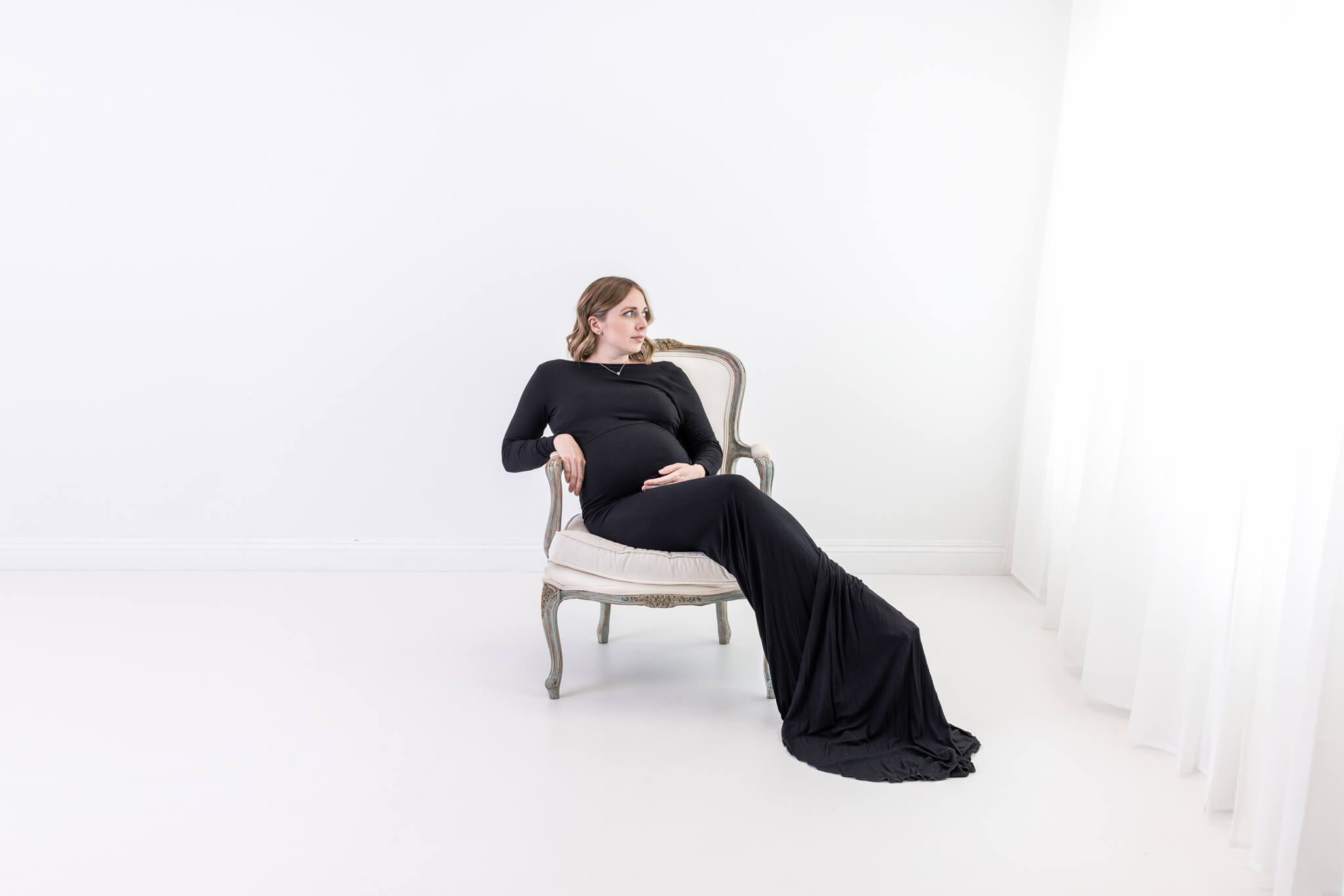 A pregnant woman in a long black dress sits on an elegant chair in a bright, minimalist room with white walls and sheer curtains, looking thoughtfully to the side.