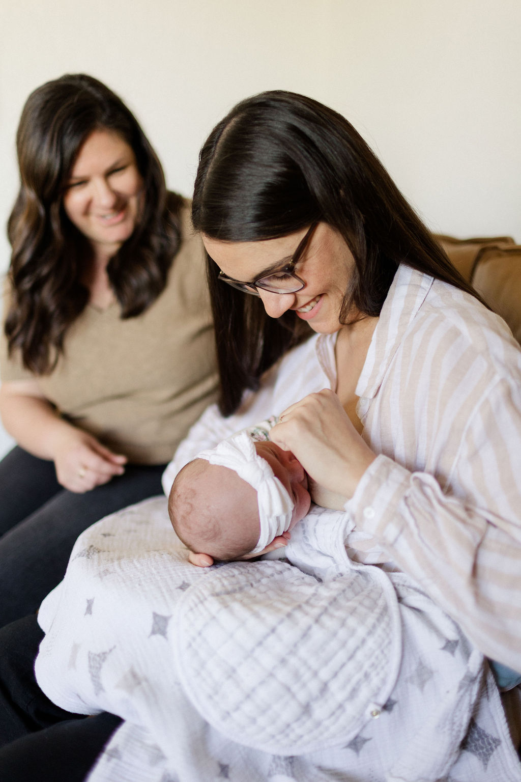 A woman at the Breastfeeding Center of Orange County wearing glasses holds and looks down at a swaddled newborn baby, while another woman sits beside them on a couch, smiling.