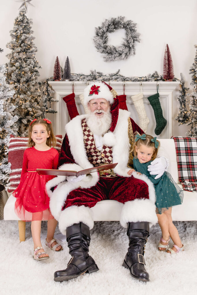 Santa Claus sits on a white bench holding a book, with two young girls in festive dresses beside him. Behind them are decorated Christmas trees, stockings, and a plaid blanket, creating a cozy holiday scene.