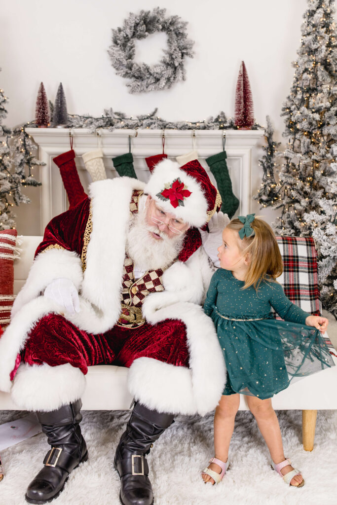 Santa Claus sits on a white bench next to a young girl in a green dress. They look at each other, surrounded by festive holiday decorations including wreaths, trees, and plaid pillows.