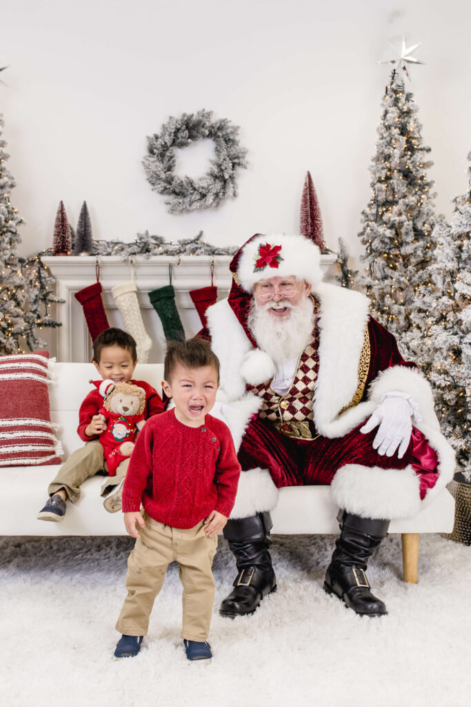 Two young children pose with Santa Claus in a festive holiday setting. One child stands in front, smiling, while the other sits on a white couch holding a teddy bear. Snowy trees and stockings decorate the background.