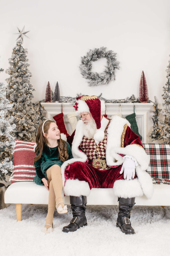 A young girl in a green dress sits on a white couch next to Santa Claus, who is dressed in his traditional red suit. They are smiling and talking, with Christmas trees and festive decorations in the background.