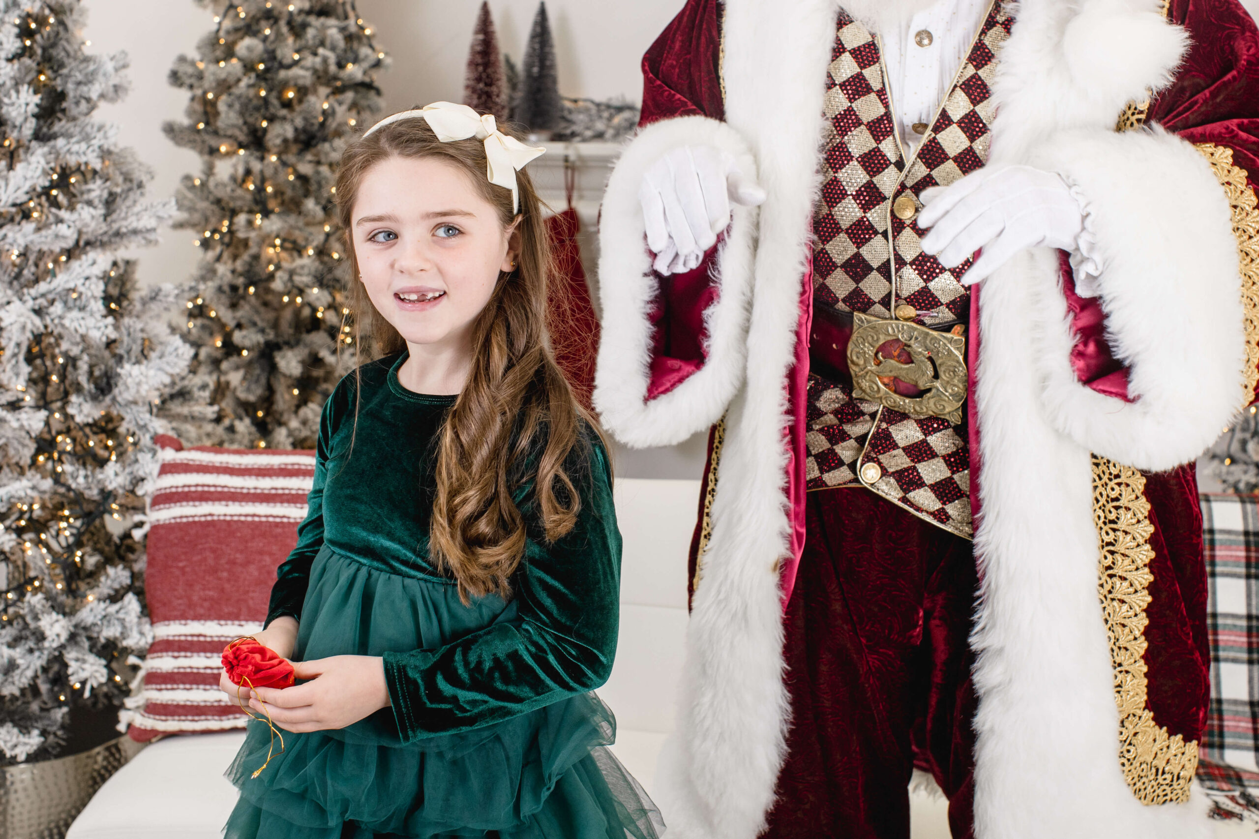 A young girl in a green velvet dress stands next to a person dressed in a Santa Claus costume, whose face is out of frame. Christmas trees and festive decorations are in the background.