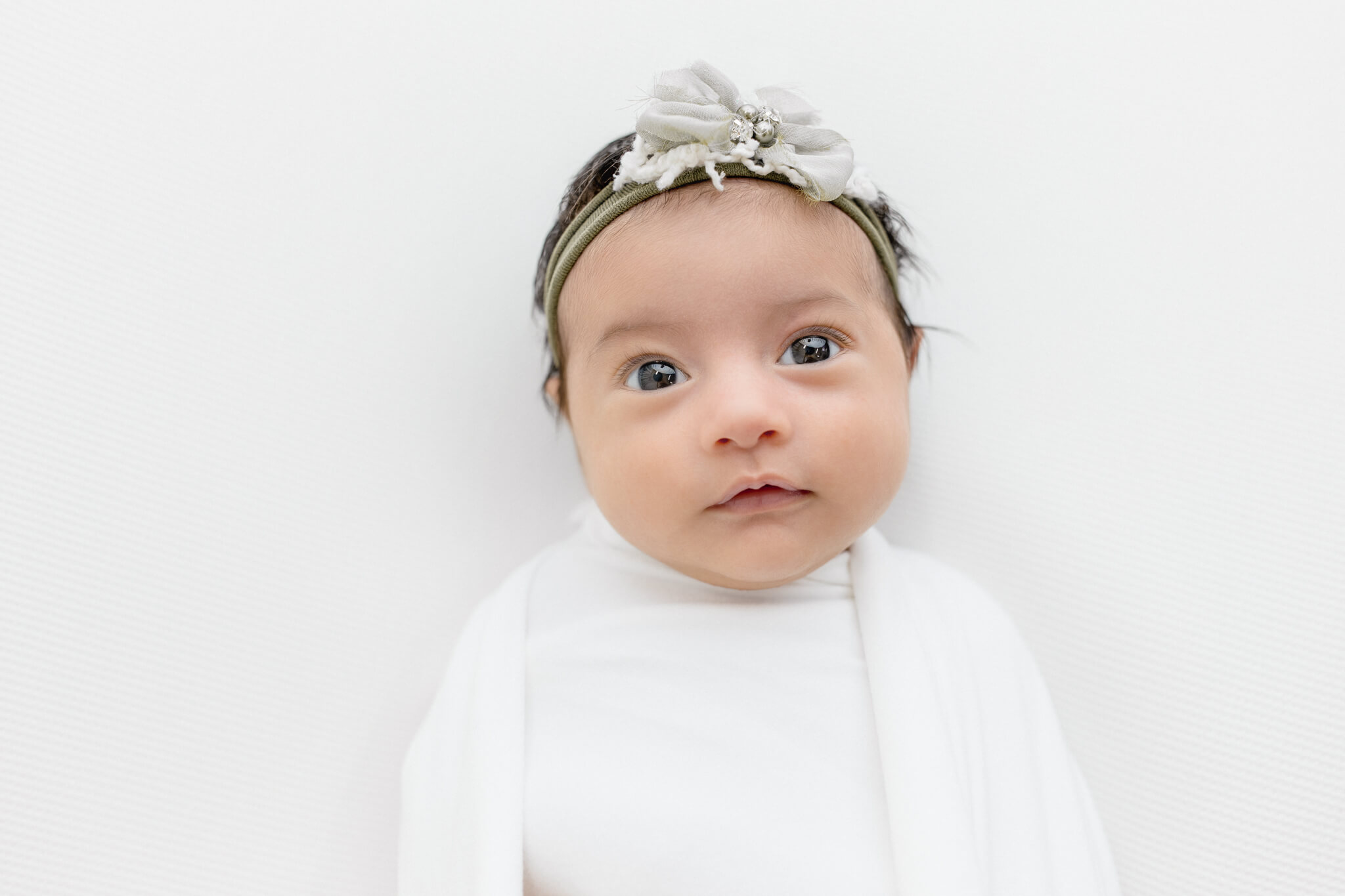 A newborn baby girl looking at the camera awake in a white swaddle wrap and headband with a bow.