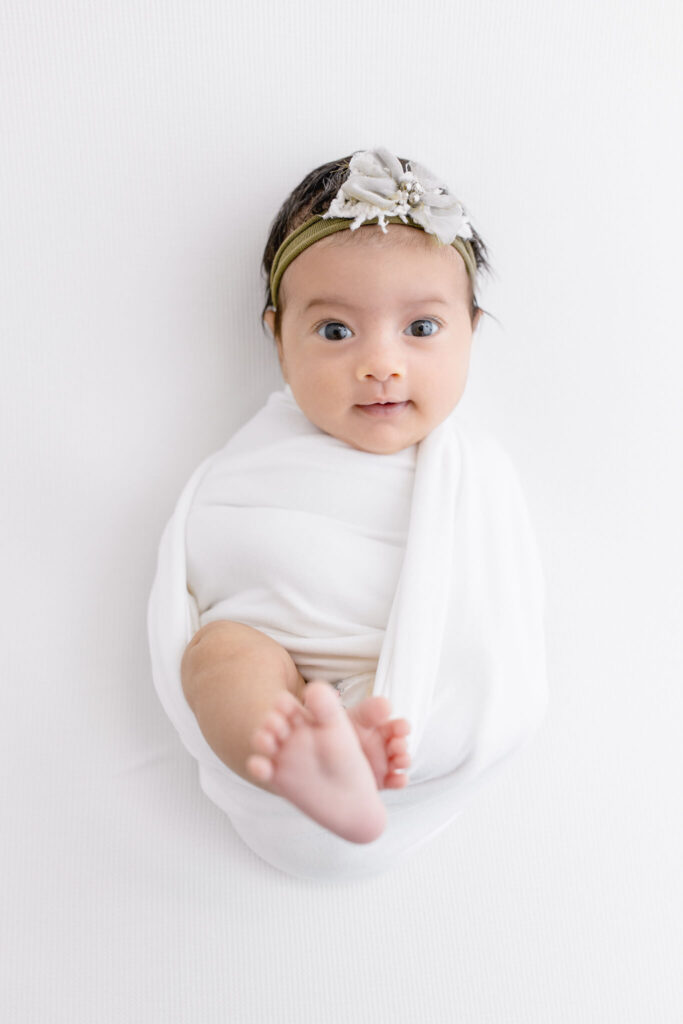 A baby wrapped in a white blanket lies on a white surface, looking up. The baby wears a green headband with a decorative bow and has one foot slightly raised toward the camera.