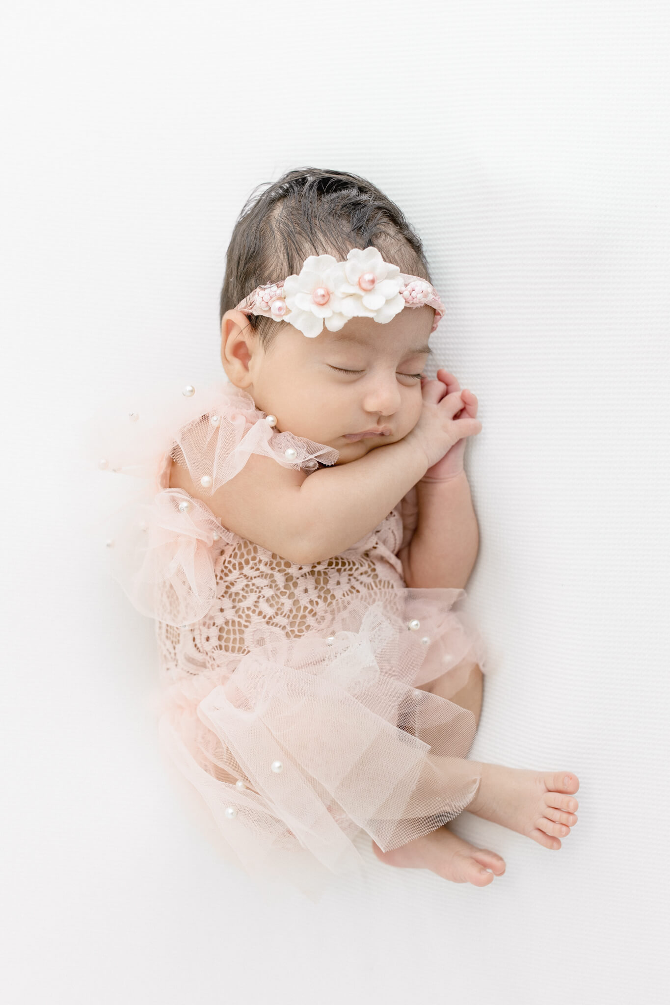 A newborn baby girl in a pink lace outfit and matching floral headband sleeps with arms curled near her face on soft white fabric